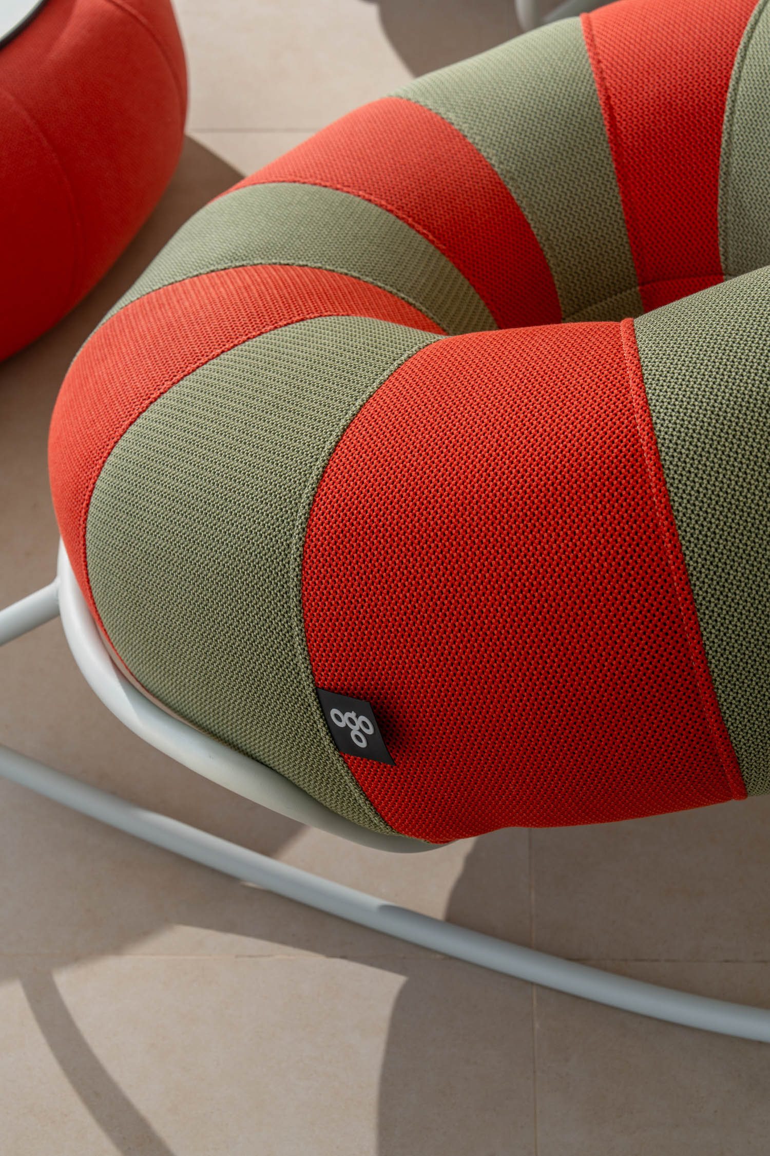 Close-up of a modern outdoor chair with red and green striped mesh fabric, featuring a small “ogo” label, on a light tile floor.