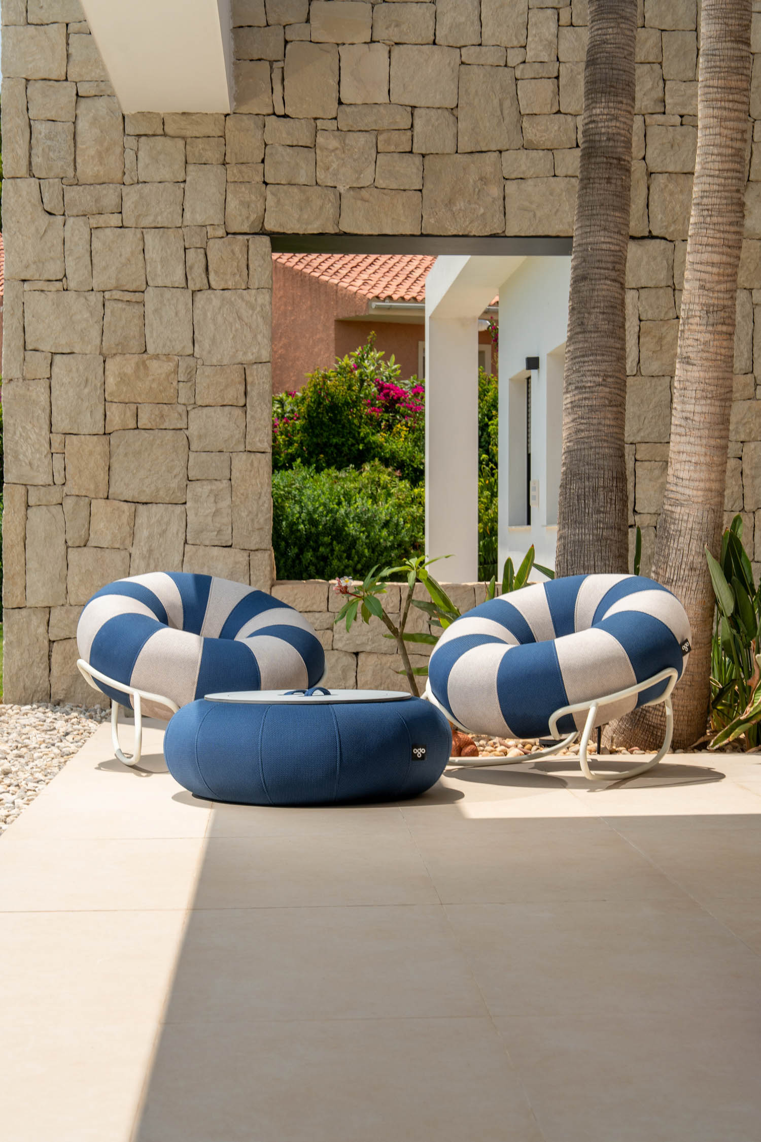 Two round, blue and white striped lounge chairs and a matching ottoman are arranged on a tiled patio with stone walls and palm trees.