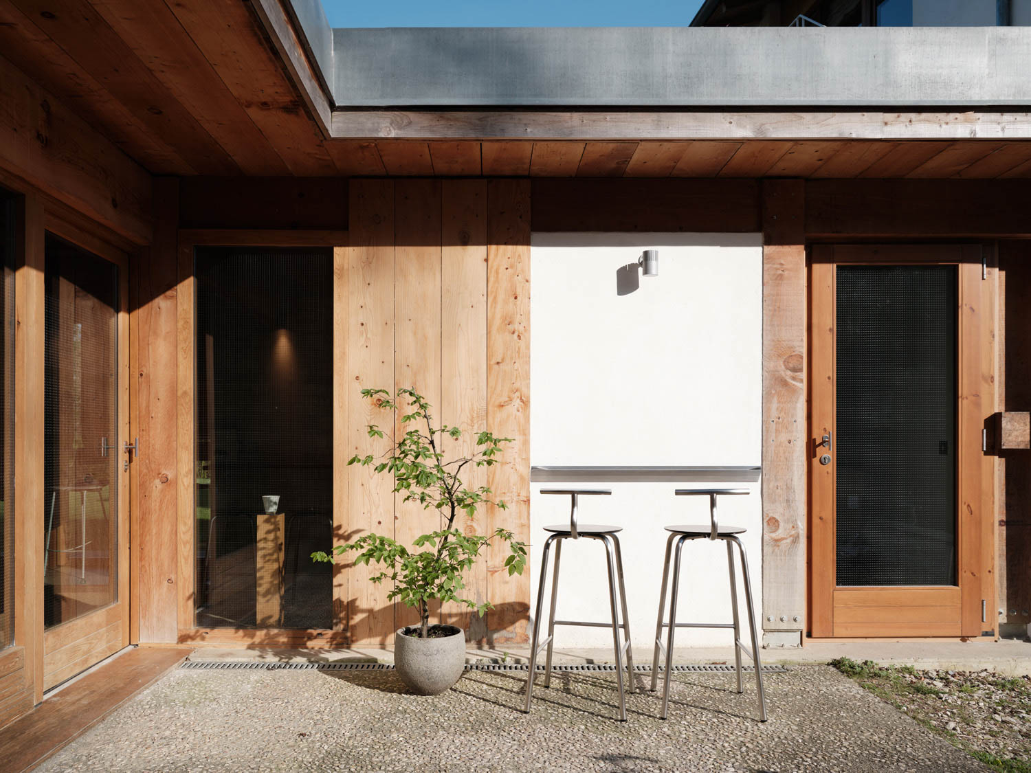 A modern courtyard with wooden walls, a potted plant, two metal bar stools, and a white panel under a small wall light.