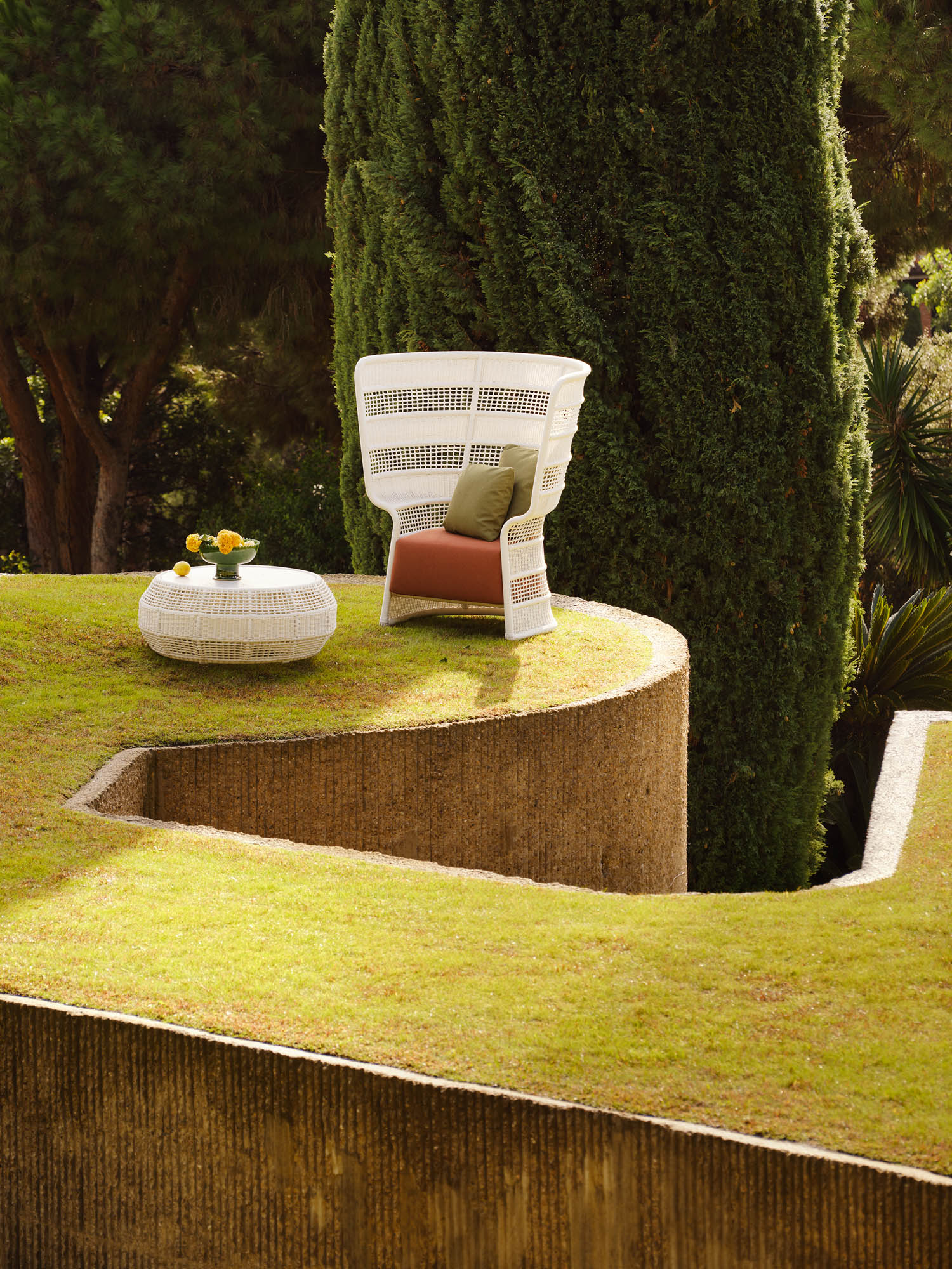 A white wicker chair with cushions and a matching table with yellow flowers sit on a grassy terrace bordered by textured stone, with tall trees in the background.
