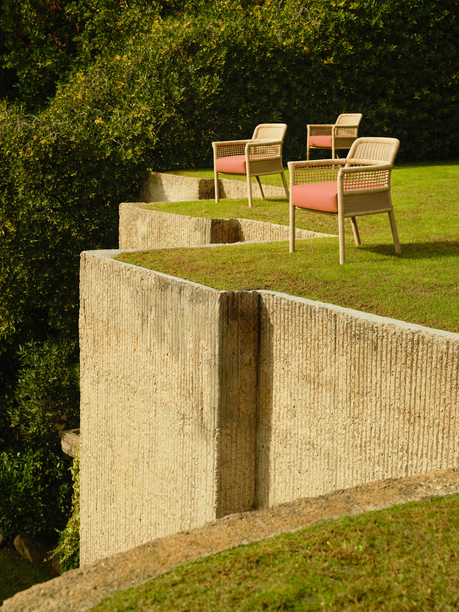 Four chairs with red cushions are arranged on a grassy terrace bordered by textured concrete walls on a sunny day, with dense greenery in the background.