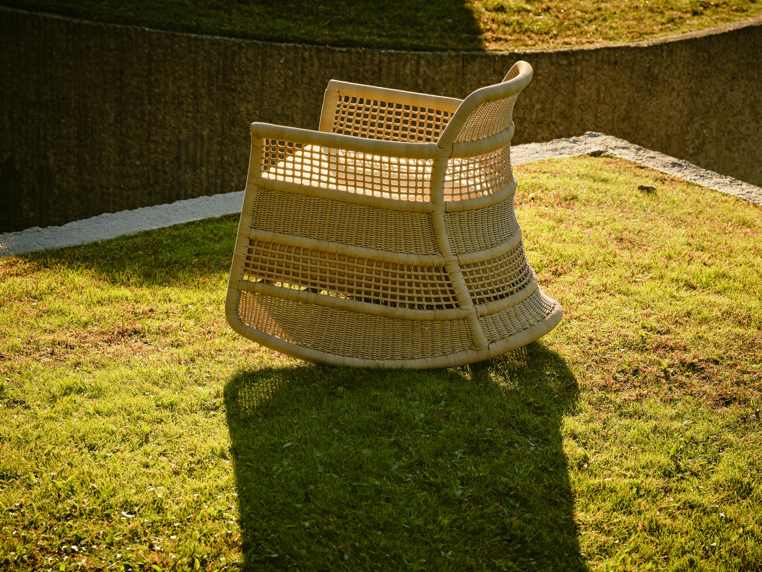 A wicker chair sits on a grassy lawn, partially lit by sunlight, with a concrete wall and shadow in the background.