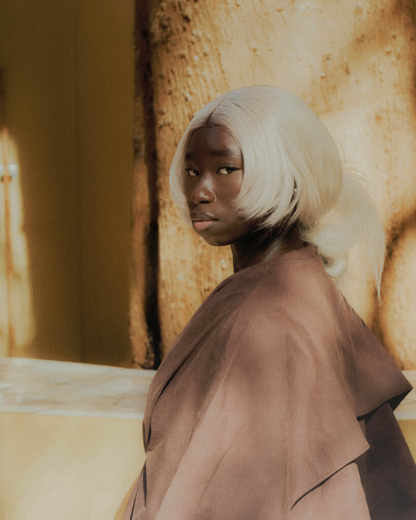 A person with platinum blonde hair and a dark brown top looks over their shoulder.