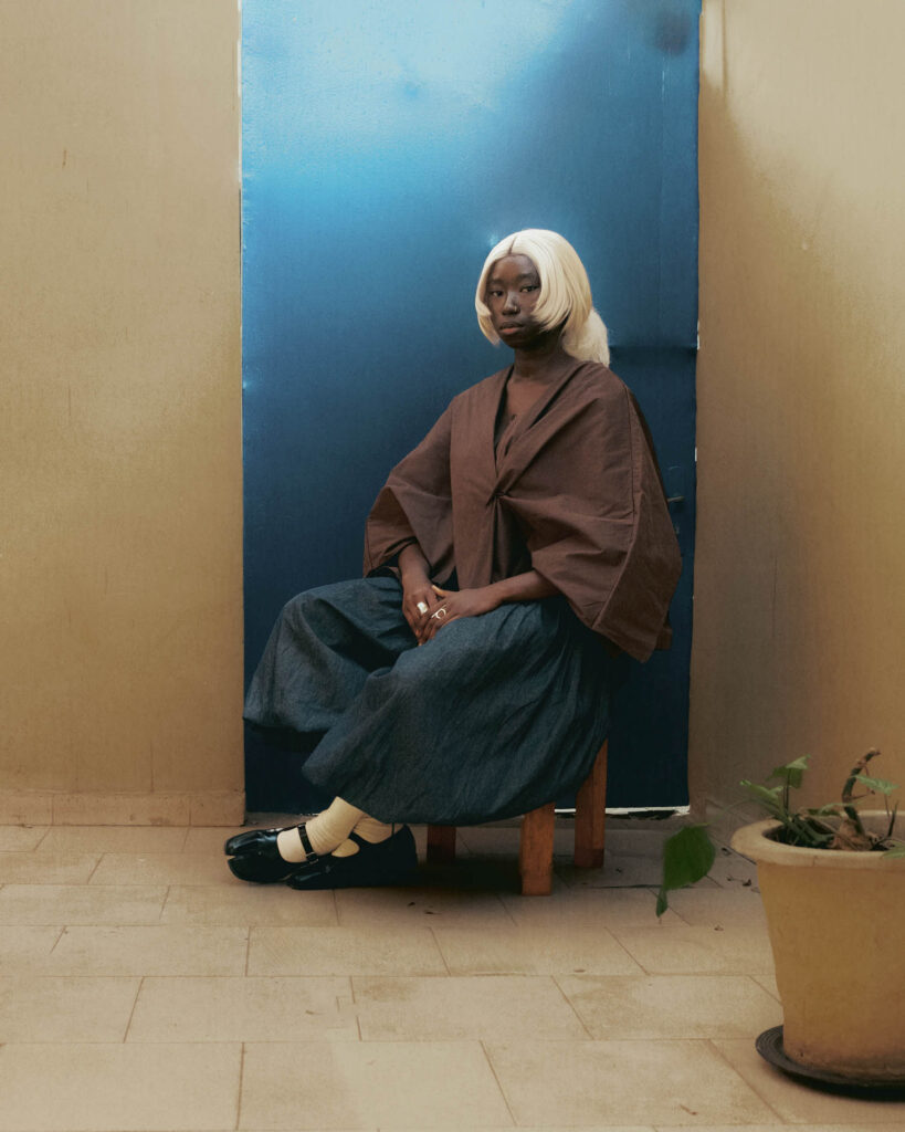 A person with platinum blonde hair and a dark brown wrap top sits on a stool in front of a blue door.