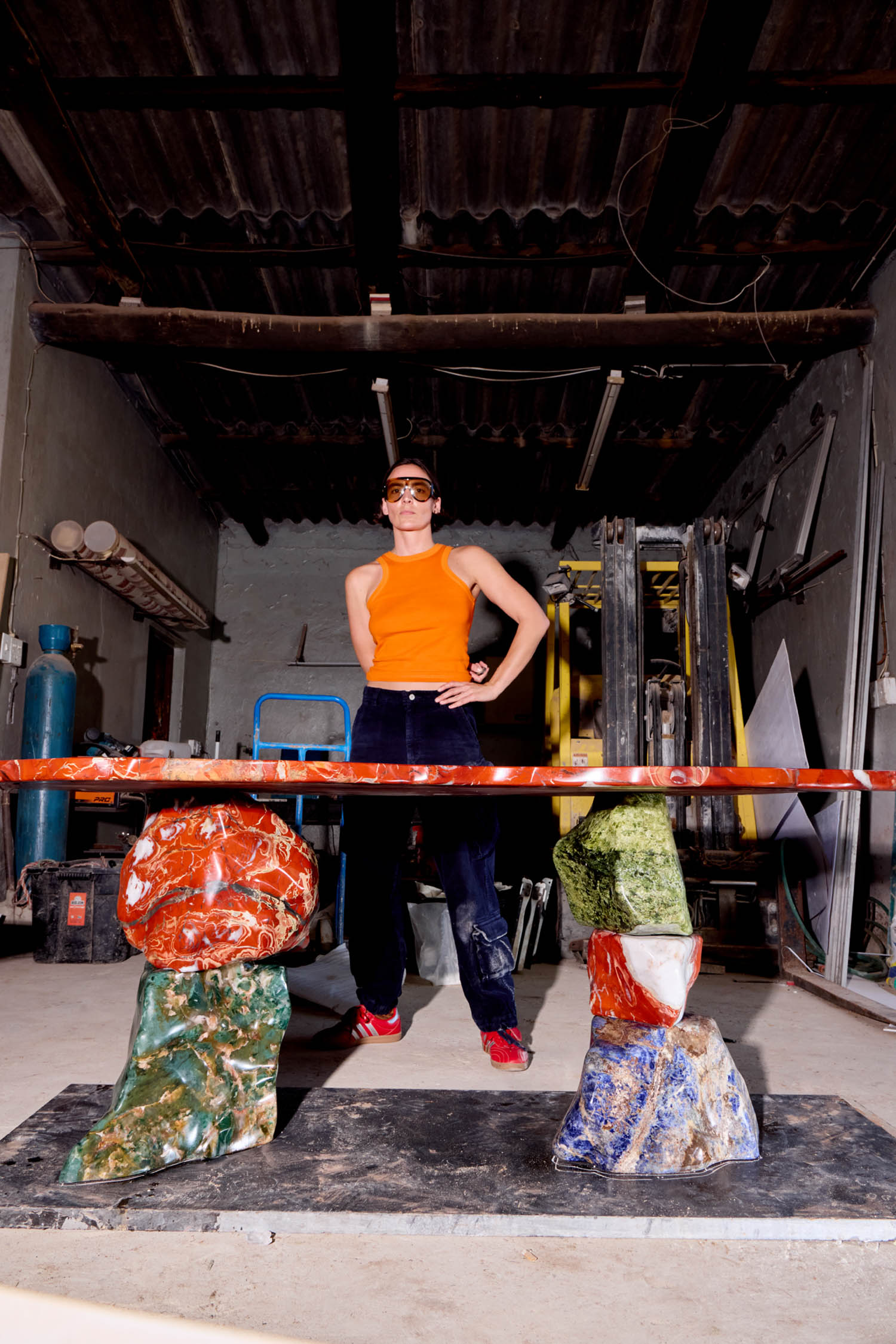 A person wearing an orange tank top and safety goggles stands behind a table supported by large, multi-colored stone bases in a workshop.