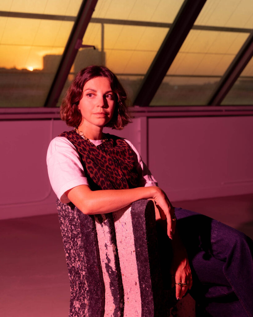 A woman with bobbed hair sits on a chair in a room with a slanted glass ceiling at sunset, bathed in red light.