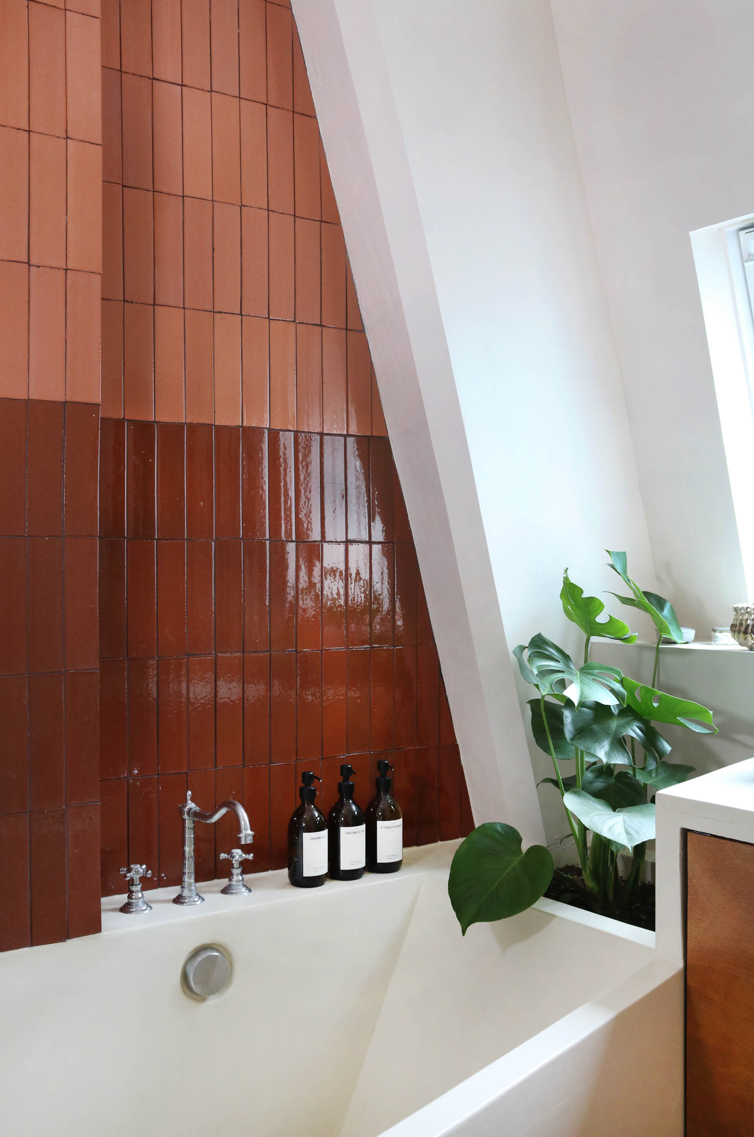 A bathtub set against a wall of dark and light brown rectangular tiles, with three amber bottles on the ledge and a potted monstera plant nearby.