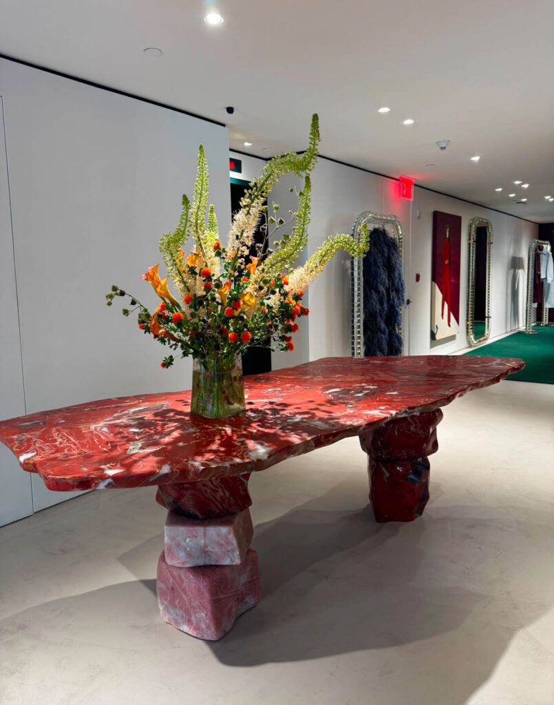 A large, irregular red marble table with sculptural block legs sits in a modern showroom, topped by a tall arrangement of orange and green flowers in a glass vase.