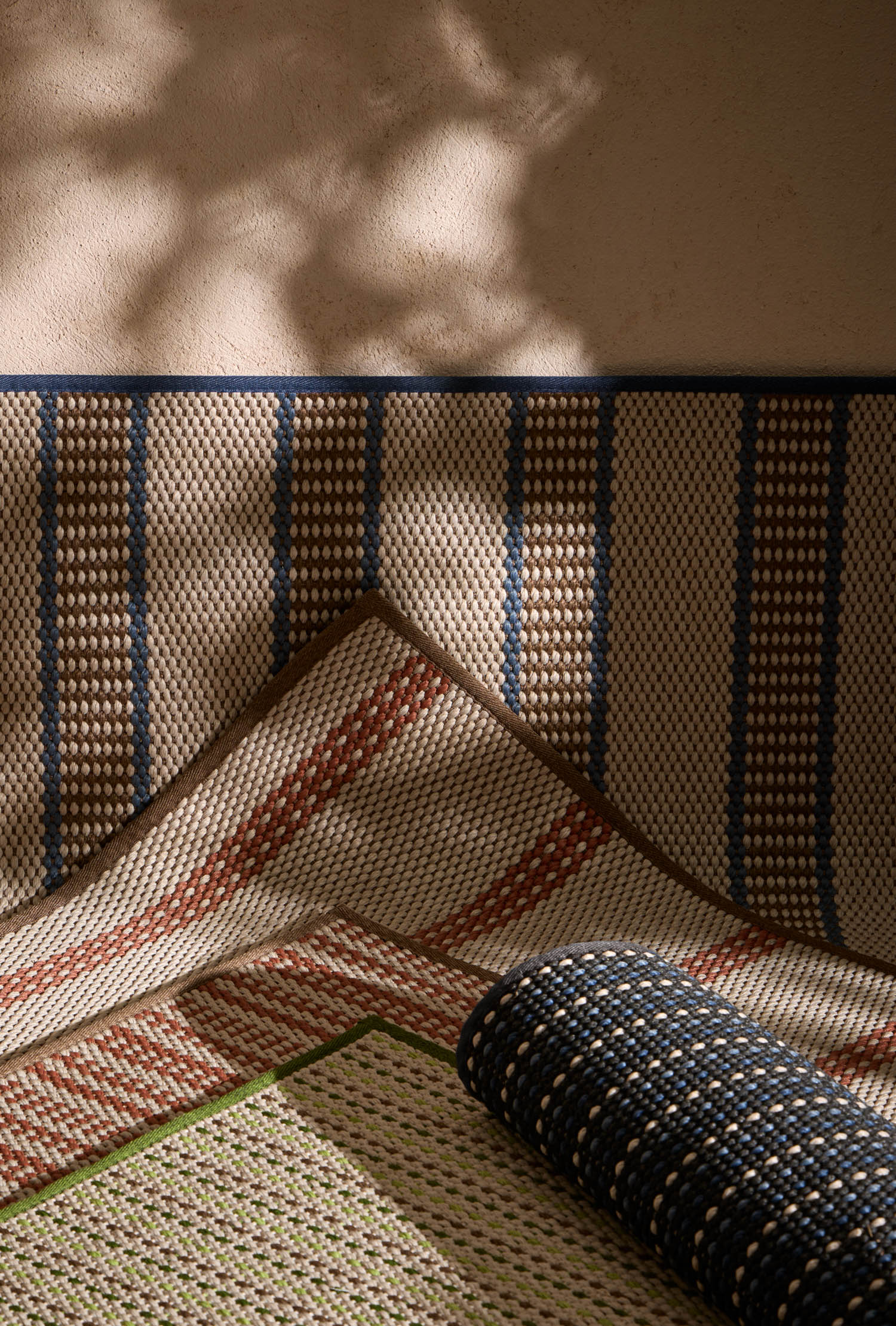 Three woven rugs with geometric patterns in blue, red, and green tones are arranged against a wall, with sunlight casting shadows on the surface.