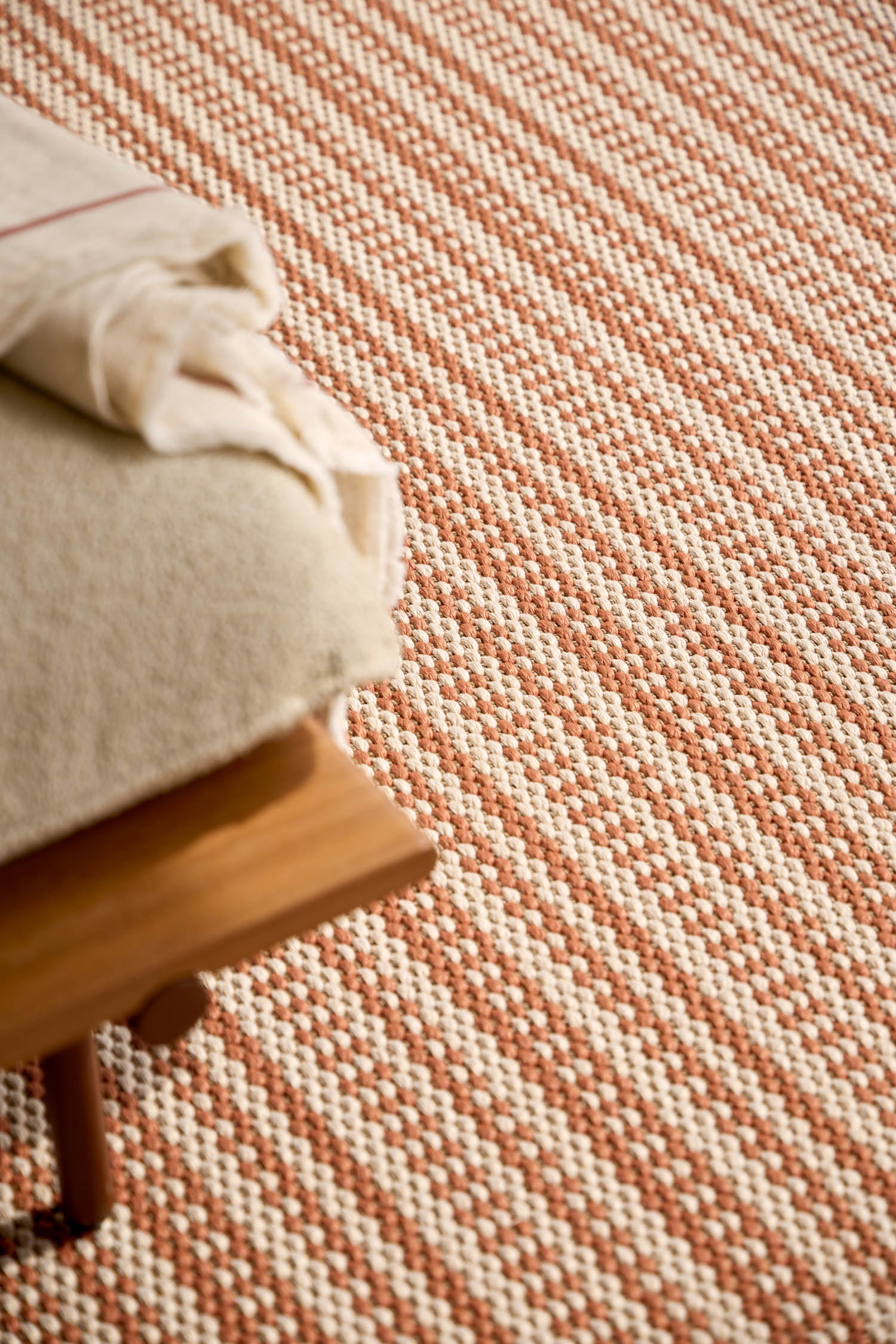 A close-up of a woven, checkered rug in brown and cream tones next to the corner of a wooden bench with folded blankets on top.