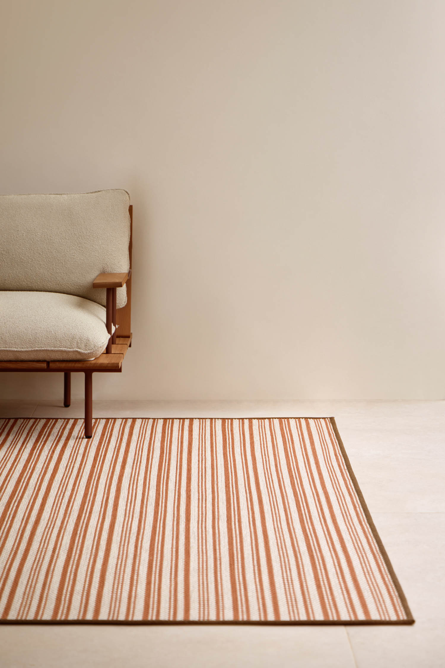 Minimalist living space with a wooden sofa and light cushions next to a beige rug featuring rust-colored stripes, set against a plain light wall.