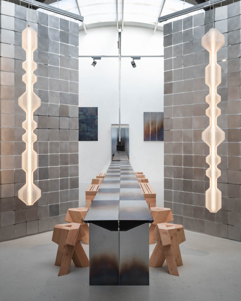 A long, thin metal table with wooden stools in a room flanked by metallic tile walls and two vertical, layered pendant lights.
