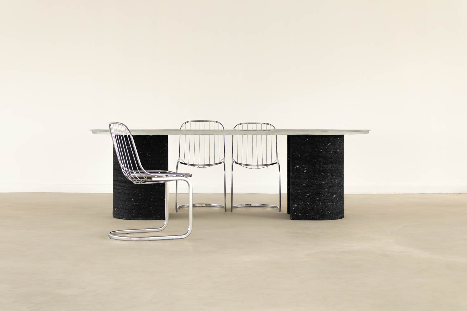 Dining table with a light-colored top and two dark, cylindrical bases, accompanied by three chrome wire chairs.