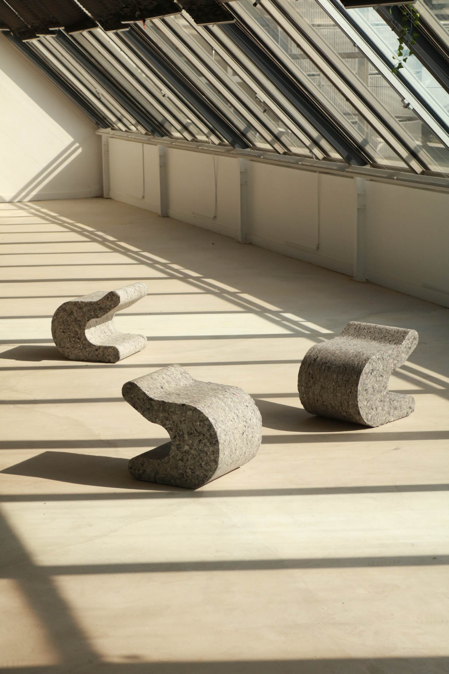 Three grey speckled, curved sculptural stools arranged on an empty floor in a sunlit room with sloped glass ceilings.