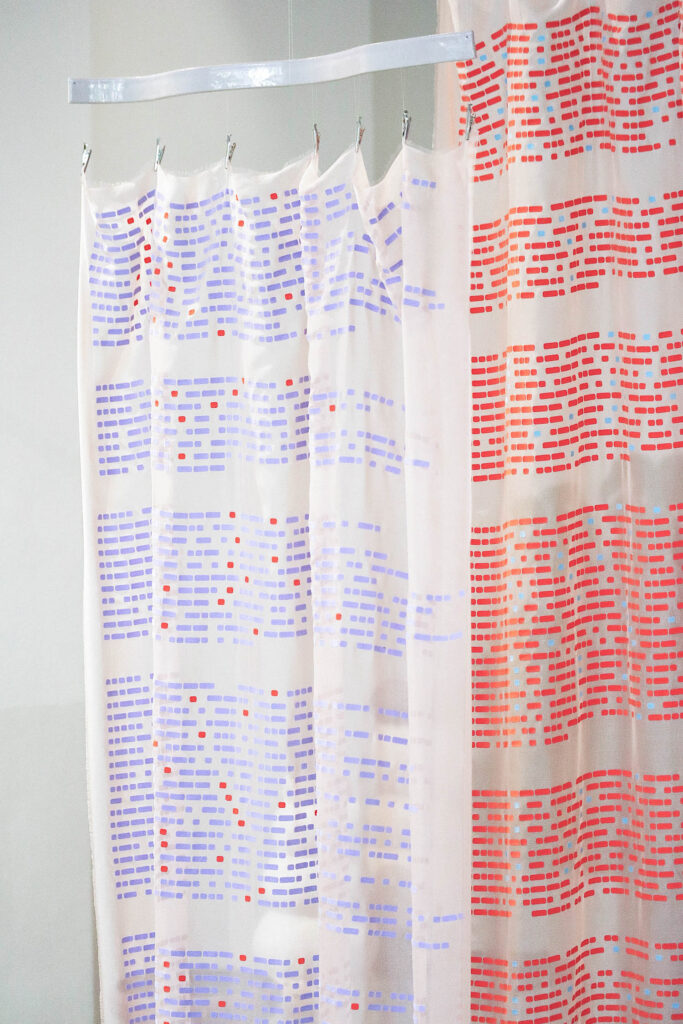 Sheer shower curtains hanging from clips, patterned with repeating blocks of purple, red, and blue dashes.