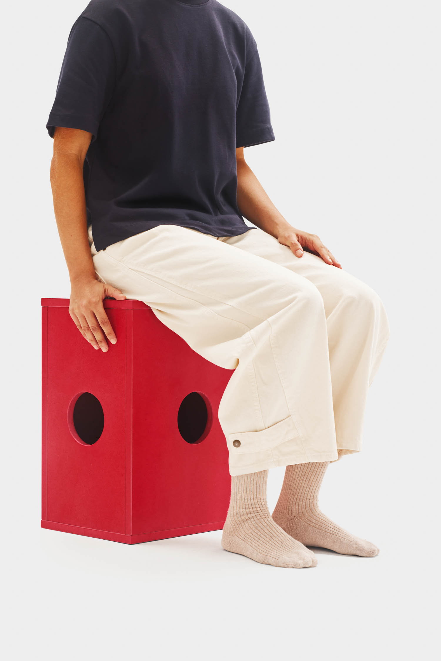 A person wearing a navy shirt, cream pants, and beige socks sits on a red box with circular holes on the side.
