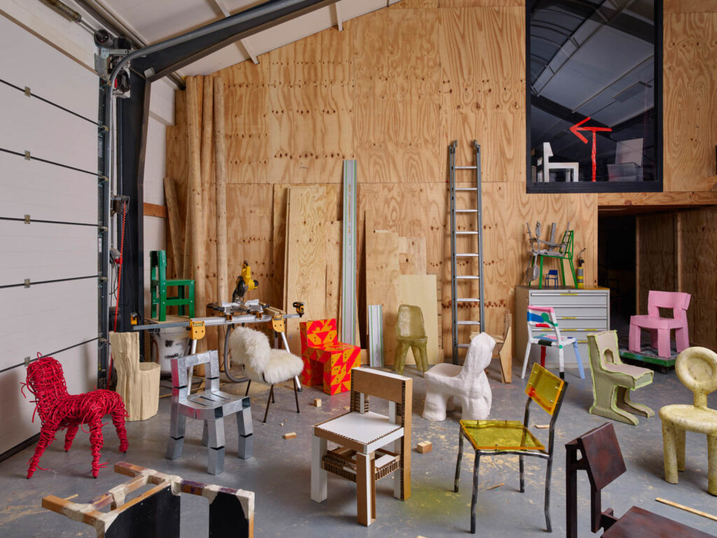 An assortment of uniquely designed artistic chairs made from various materials in a plywood-walled workshop.
