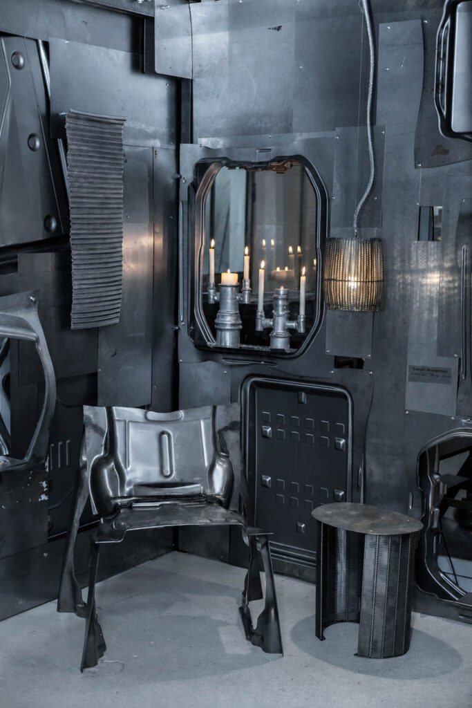 A room with walls made of metal panels, featuring a metal chair, a cylindrical side table, a hanging cage light, and a wall-mounted mirror reflecting lit candles.