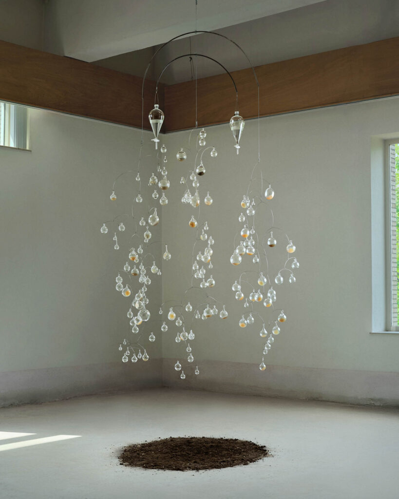 A kinetic sculpture of glass orbs suspended from wires hangs above a mound of soil on a gallery floor.