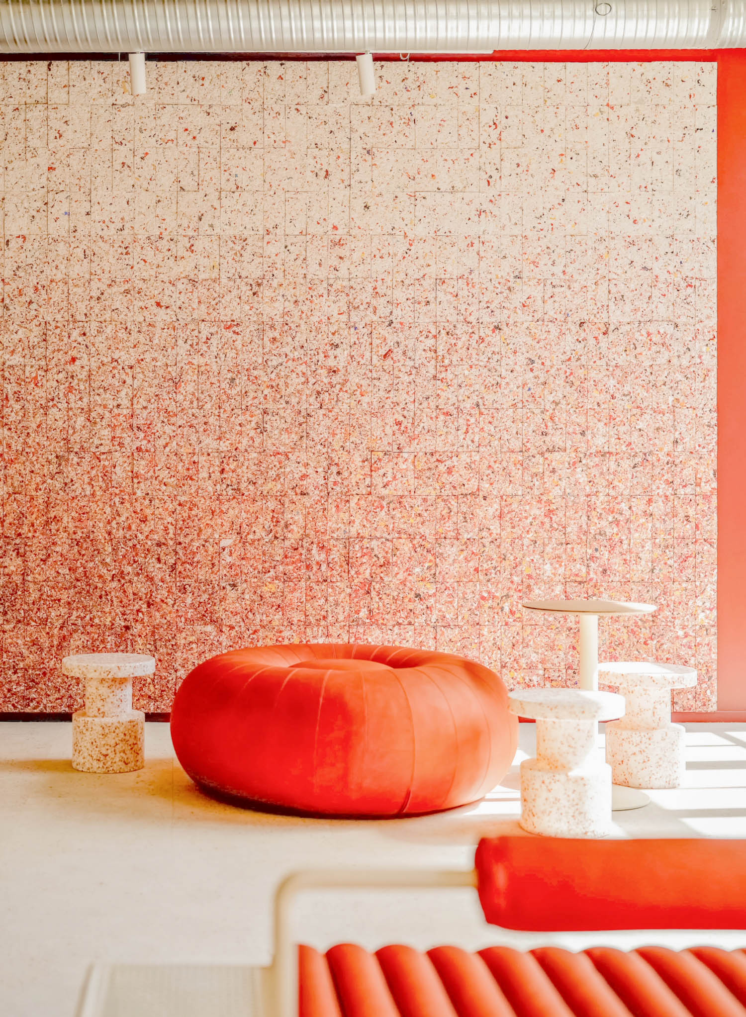 A large orange floor cushion, three small speckled stools, and a side table set against a wall with a speckled red and white tiled pattern.