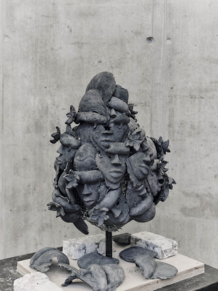 A dark, clay sculpture of multiple human faces and flowers clustered together on a wire frame, mounted on a wooden base.