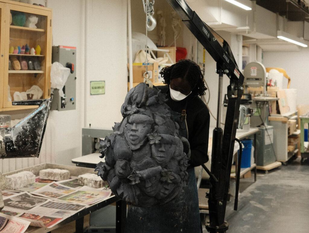 A person wearing a face mask uses a shop crane to lift a large, dark, textured sculpture of multiple faces.