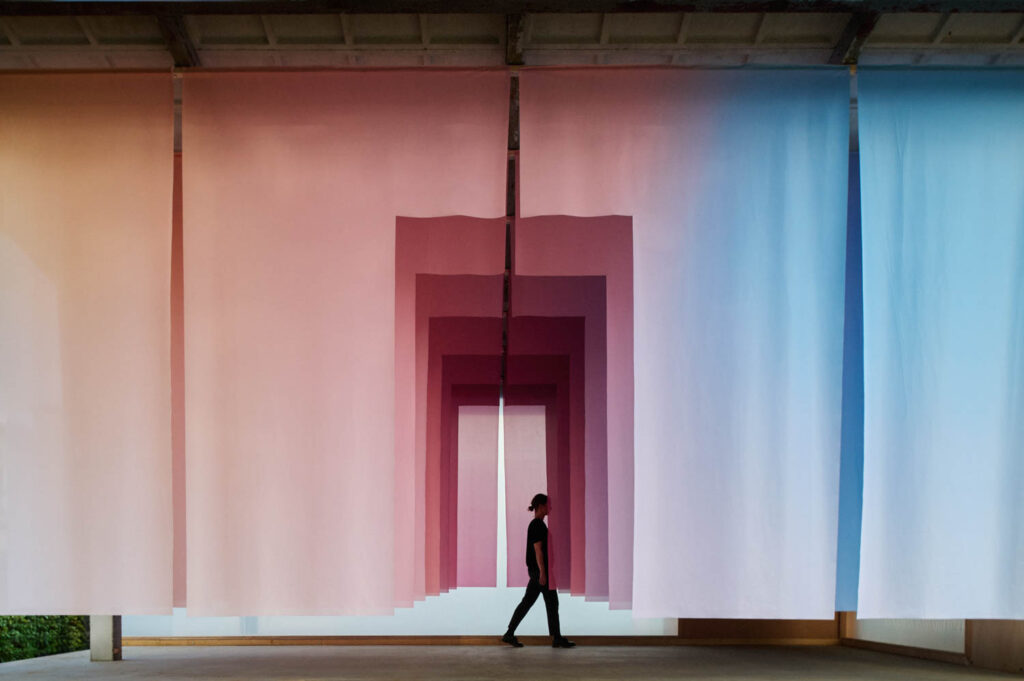 A person walking through a series of hanging fabric panels that create a pink and blue gradient tunnel.