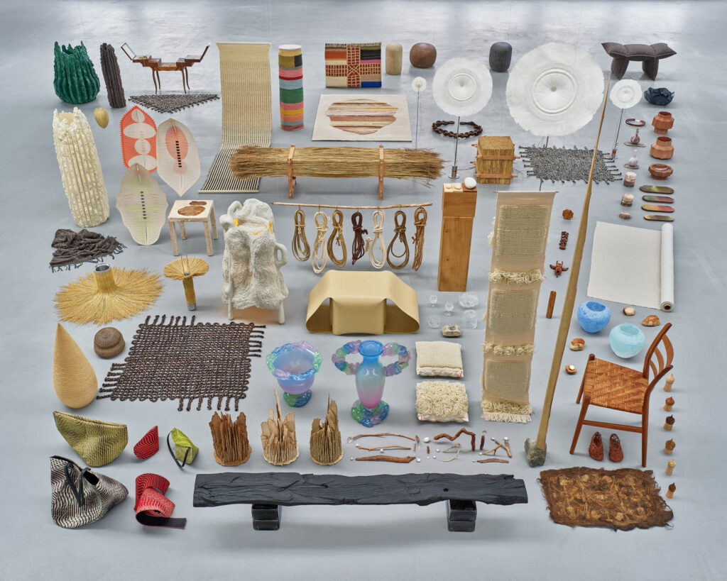 An array of artisanal objects, including woven textiles, pottery, furniture, and sculptural pieces, arranged on a gray floor.