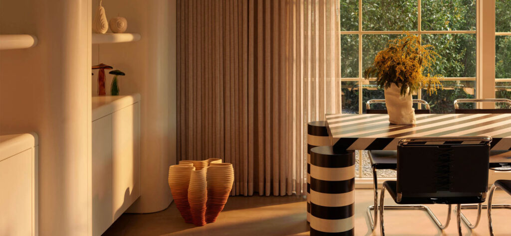 A room featuring a dining table with a black-and-white striped base and top, a sculptural gradient-colored floor vase, and a vase of yellow flowers on the table.