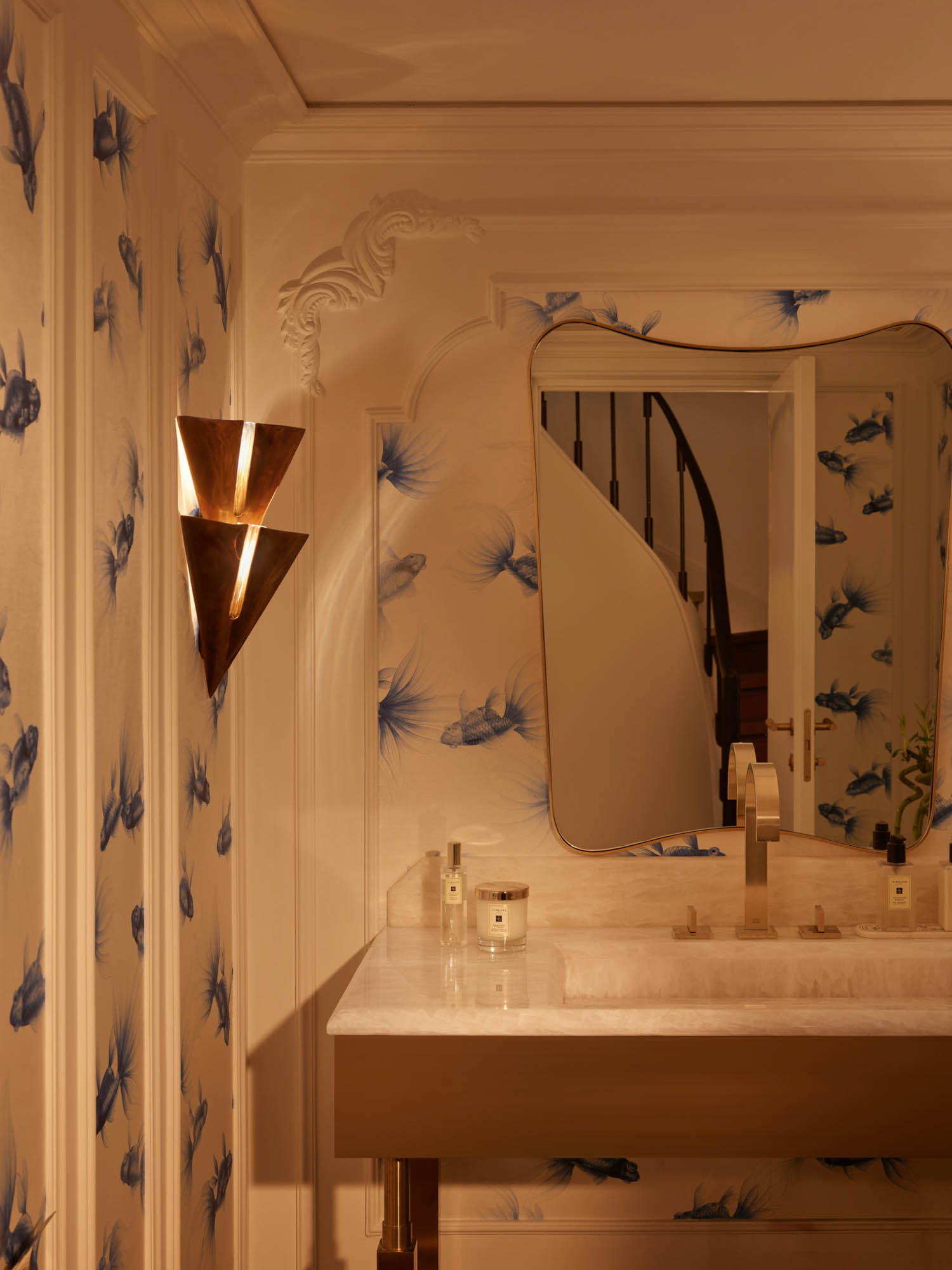 A bathroom vanity with a stone countertop, an arched mirror, and brass sconces, set against wallpaper featuring blue fish illustrations.
