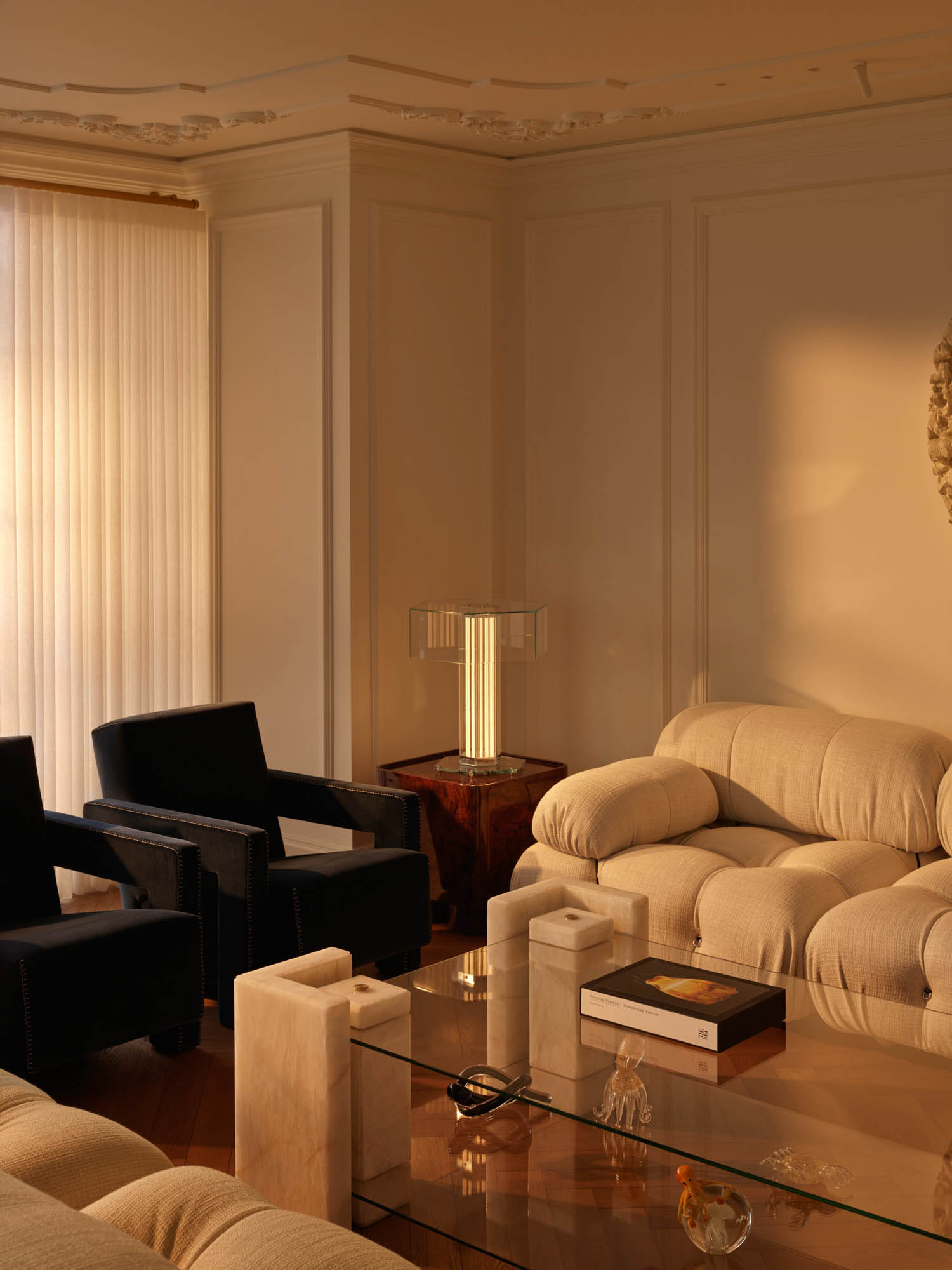 A living room with two dark armchairs, a beige sofa, and a glass-topped coffee table with stone supports, illuminated by a table lamp.