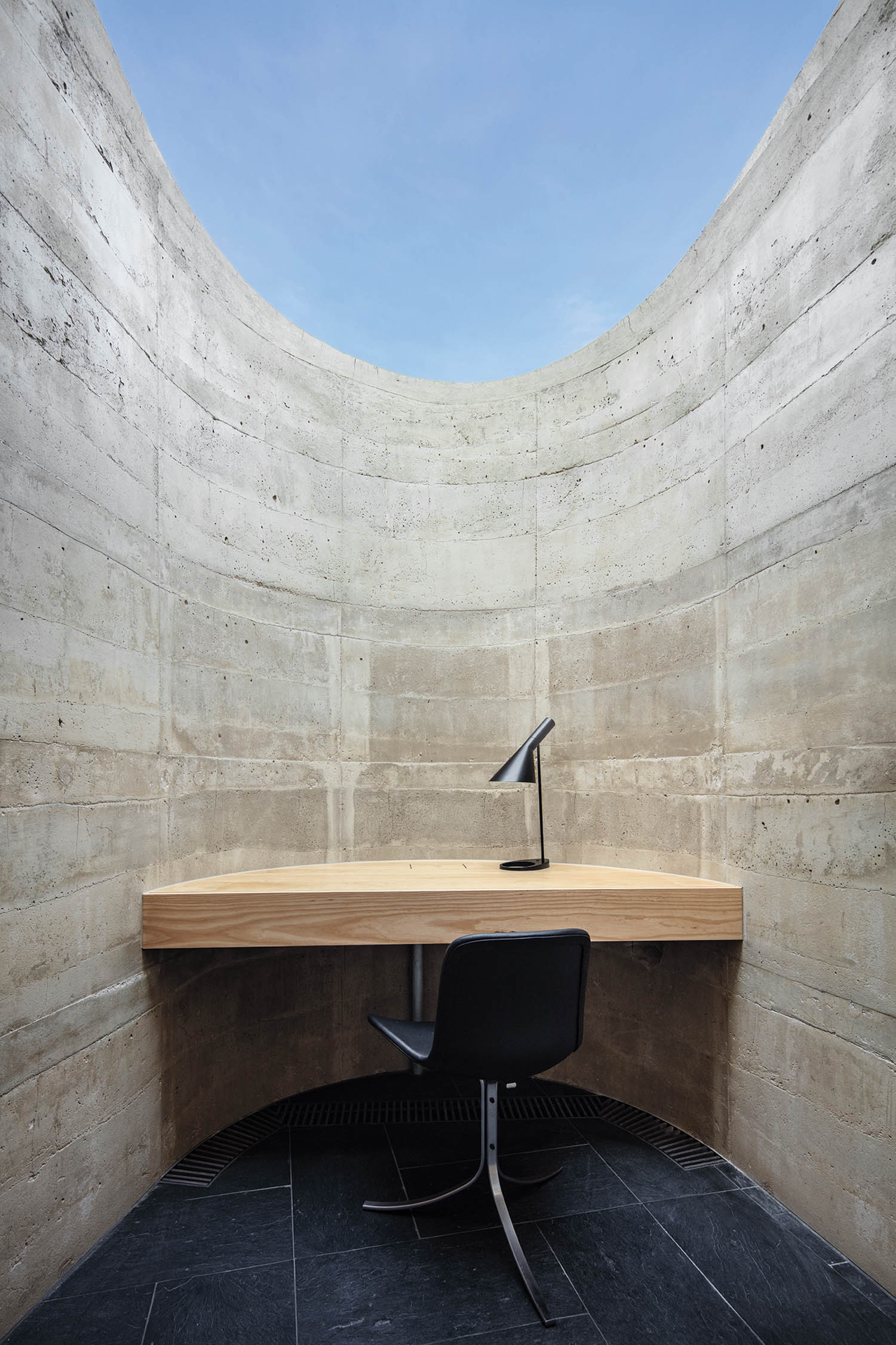 A desk and black chair set against a curved concrete wall, open to the sky above.
