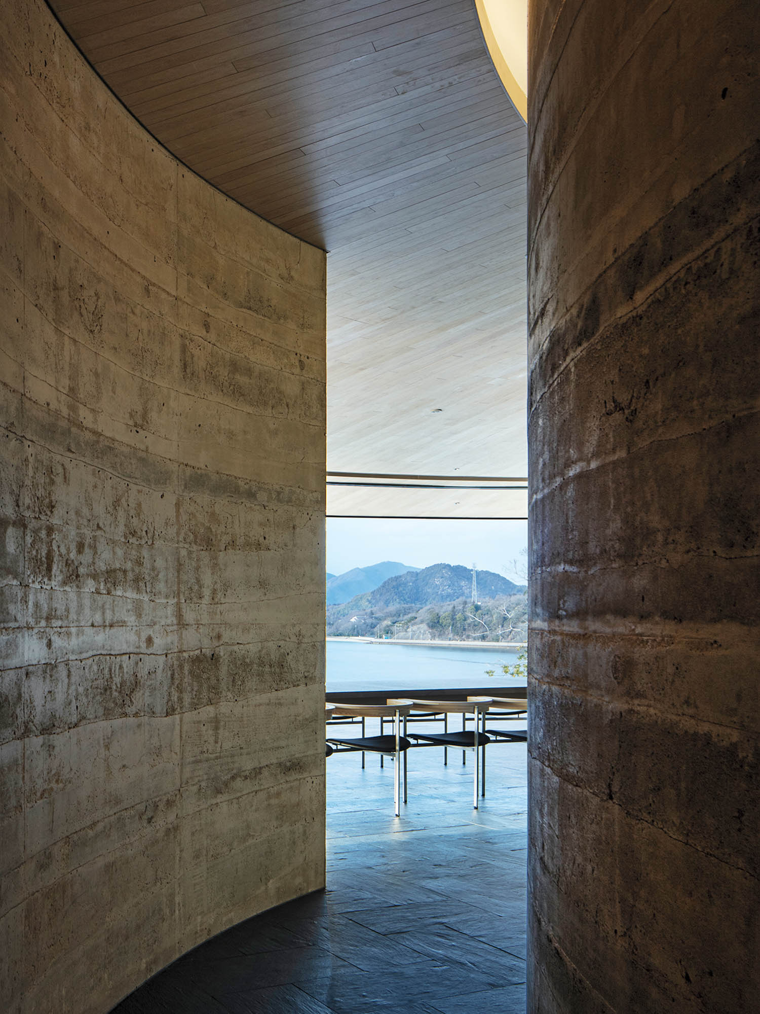 A view through a narrow gap between curved concrete walls toward a modern dining area overlooking a lake and mountains.