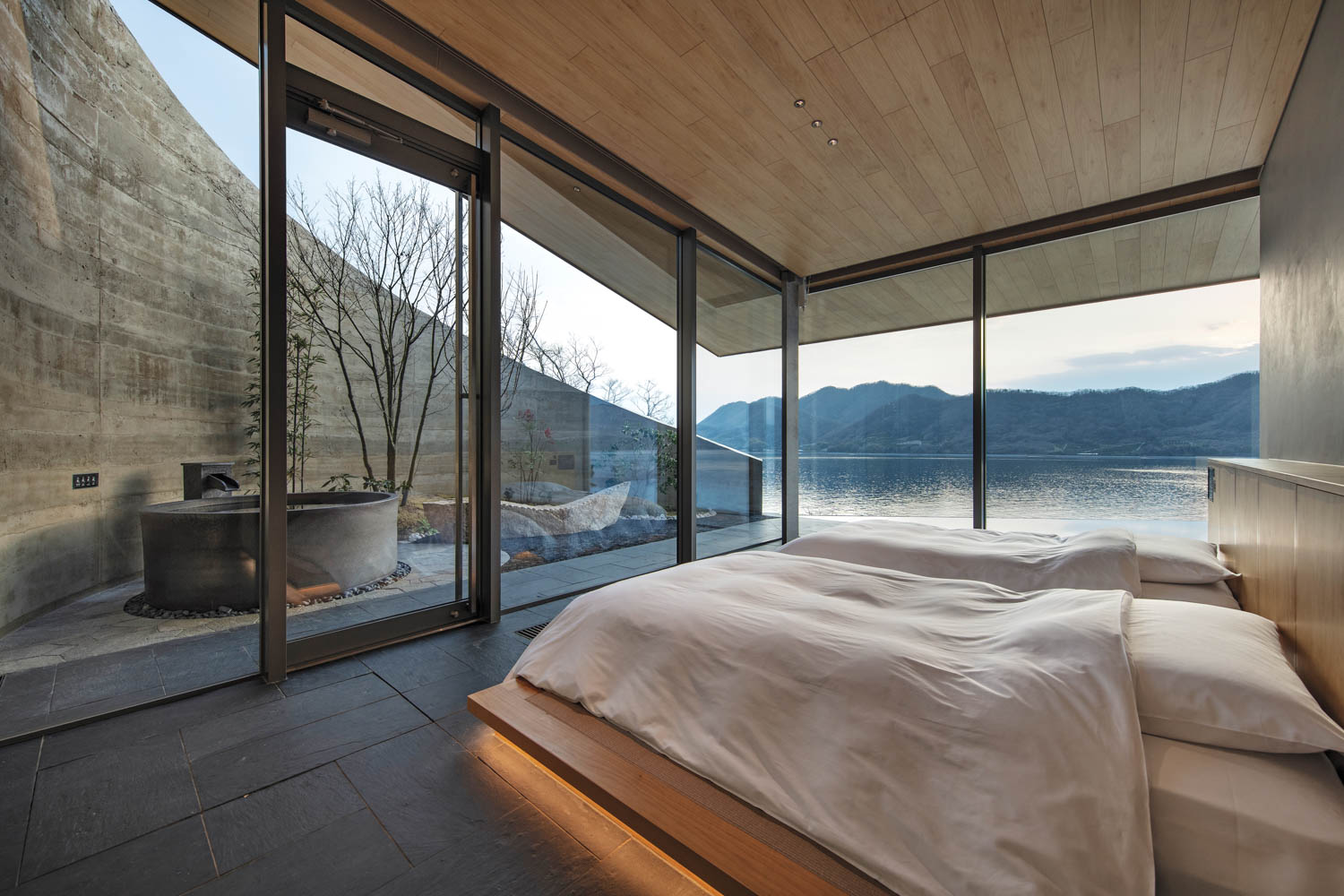Bedroom with two beds, floor-to-ceiling windows overlooking a lake and mountains, and an outdoor soaking tub on a private terrace.