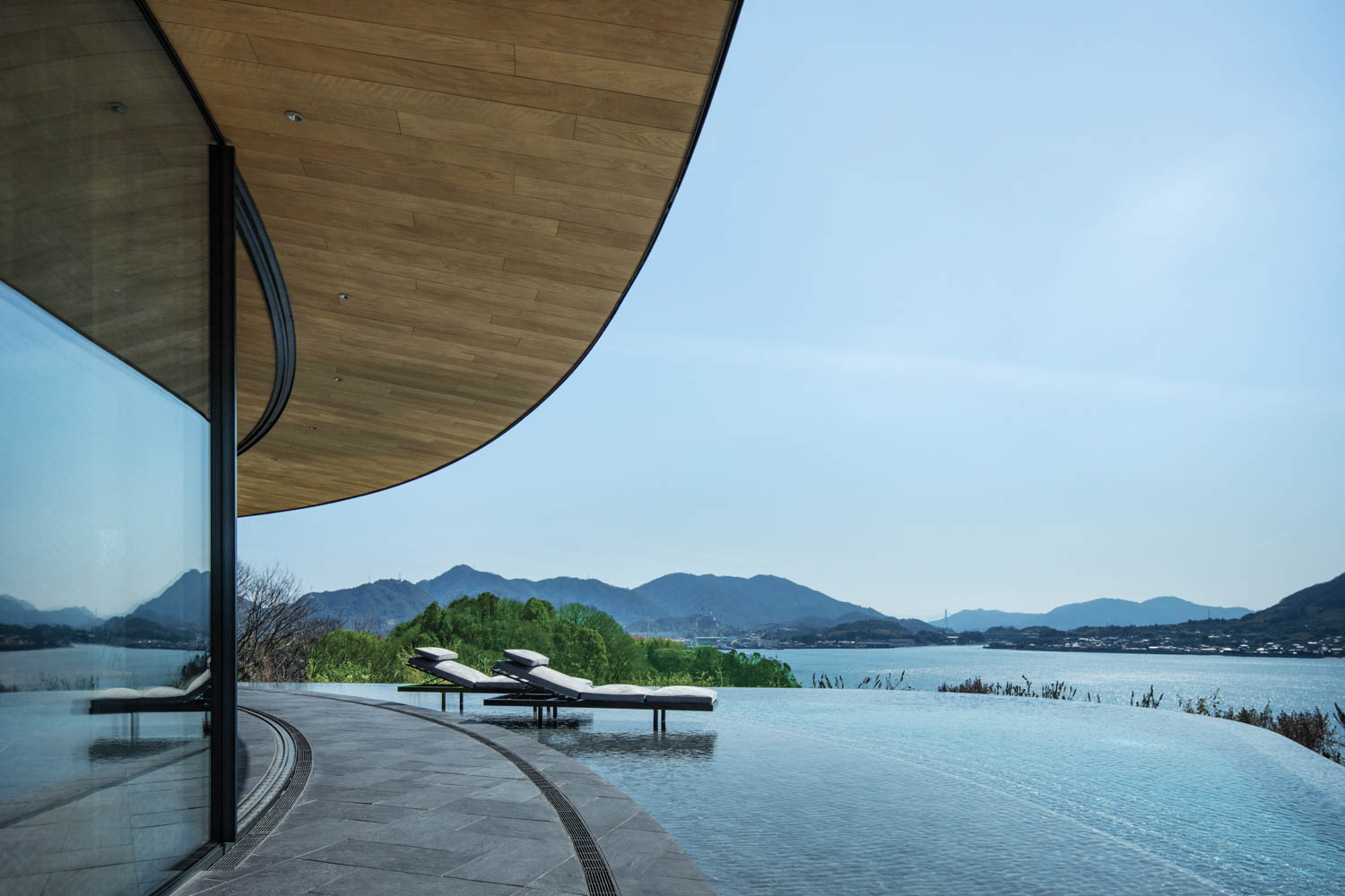 Two lounge chairs sit on a patio beside an infinity pool overlooking a bay and mountains.