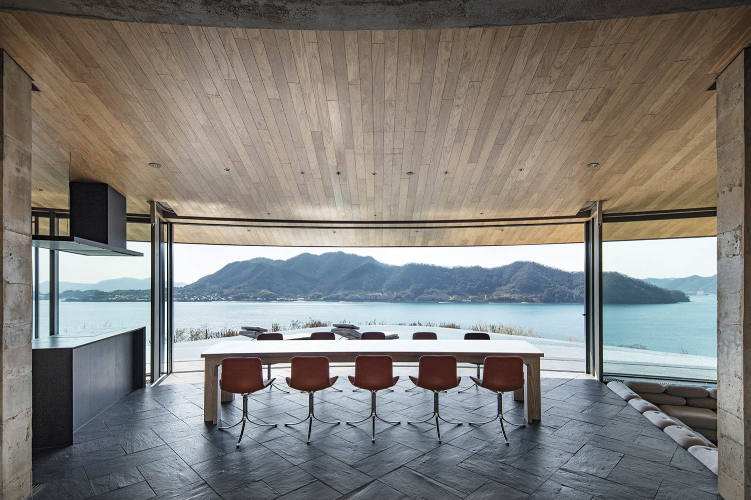A long dining table with six chairs facing a floor-to-ceiling view of a lake and mountains.