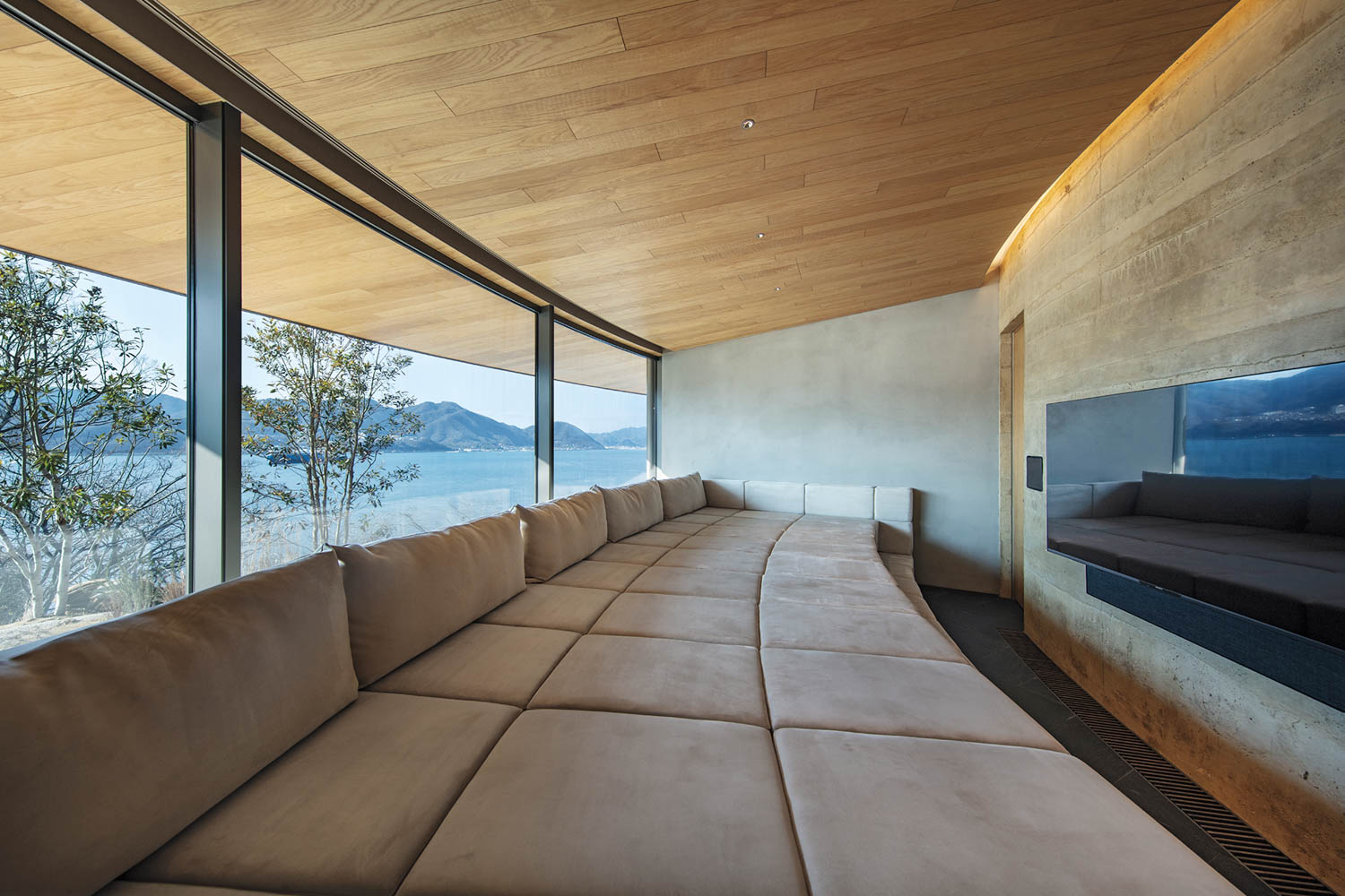 A modern living area with a long, built-in sectional sofa facing floor-to-ceiling windows overlooking a lake and mountains.