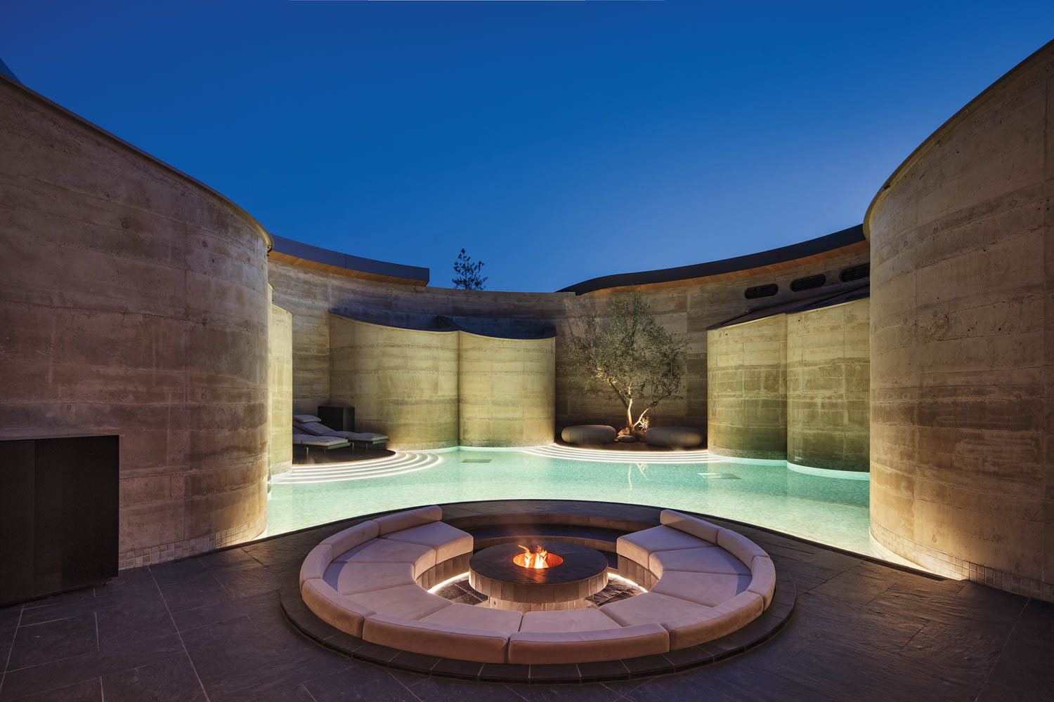 A sunken circular seating area with a central fire pit sits in the middle of a swimming pool surrounded by curved concrete walls at dusk.