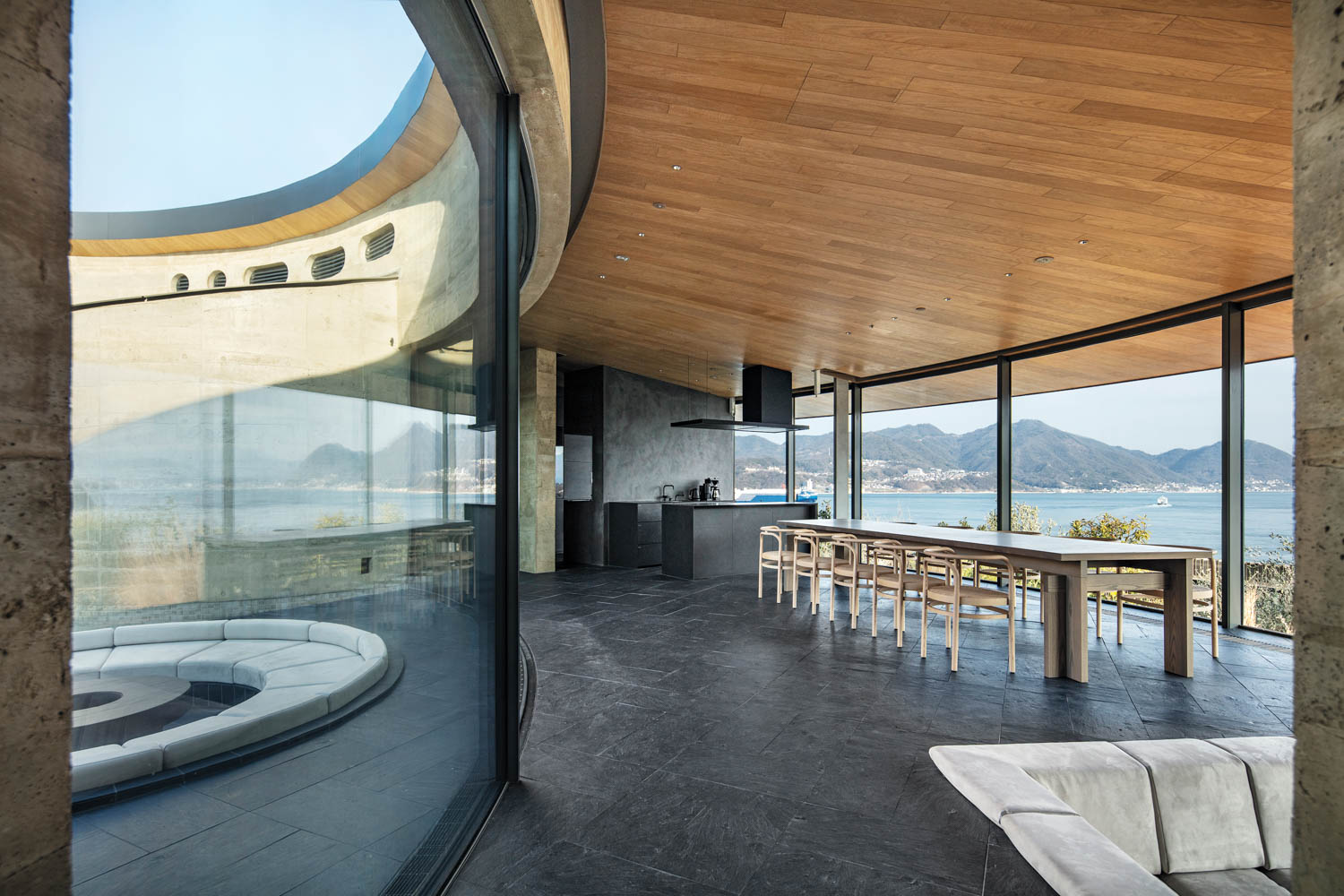 Modern open-plan interior with a large wooden dining table, a curved sunken seating area behind glass walls, and an expansive view of the ocean and mountains.