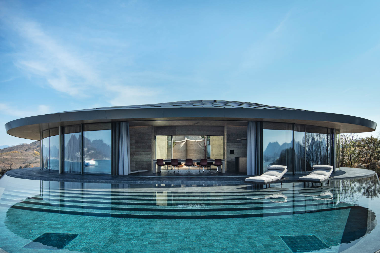 A circular modern house with floor-to-ceiling glass walls, opening onto an infinity pool with two lounge chairs on the deck.