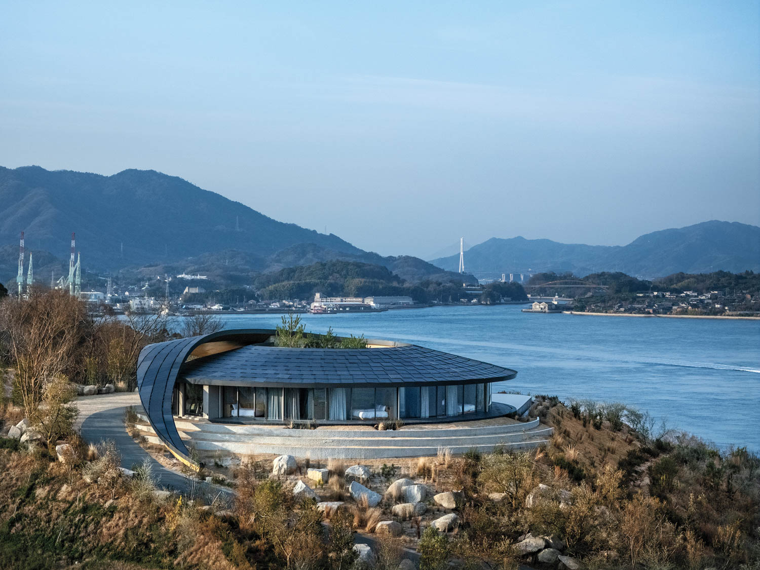 A circular house with a sweeping, curved roof overlooking a bay and mountains.