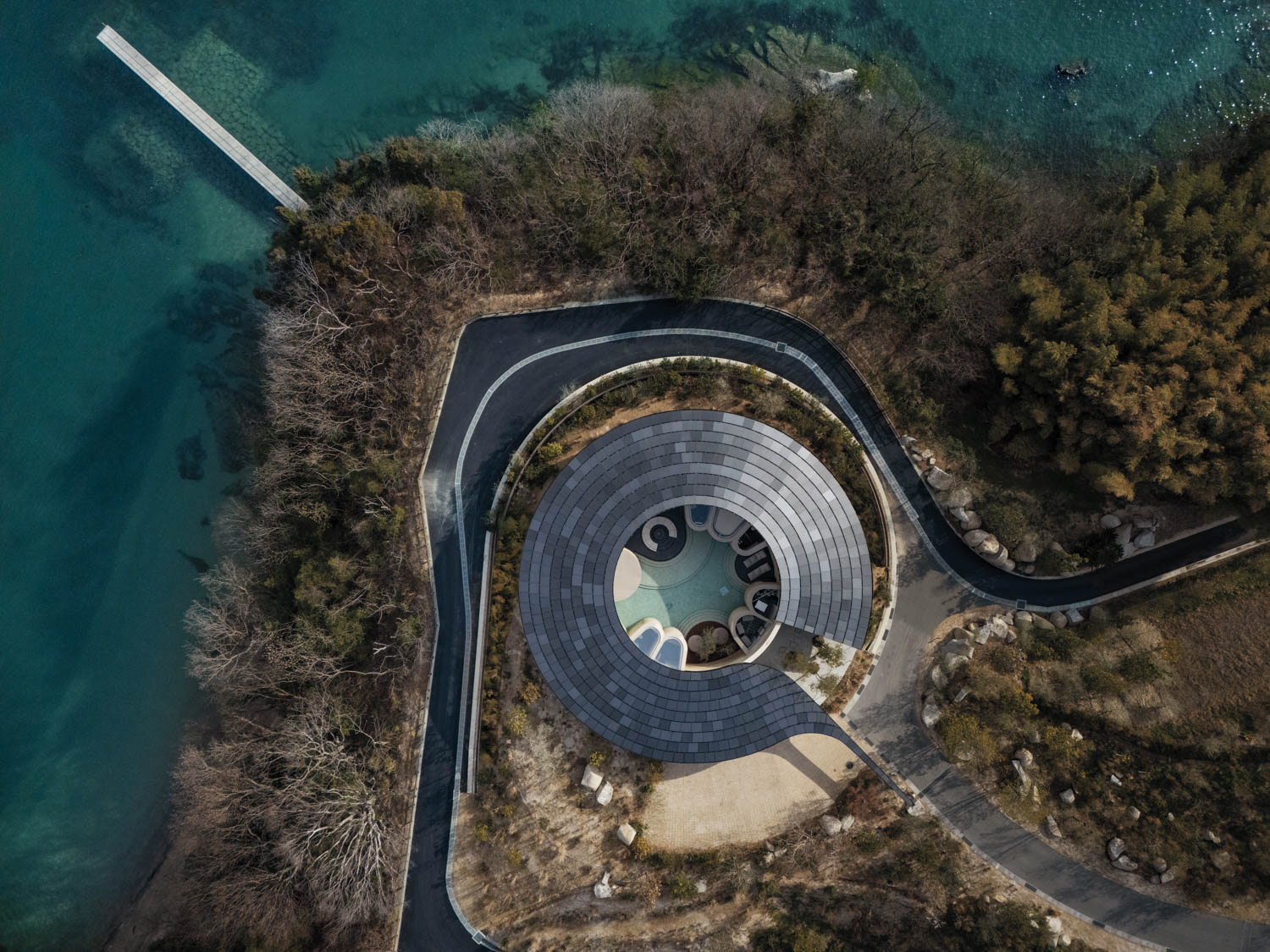 Aerial view of a circular building with a curved roof located on a wooded coastal peninsula with a pier extending into the water.