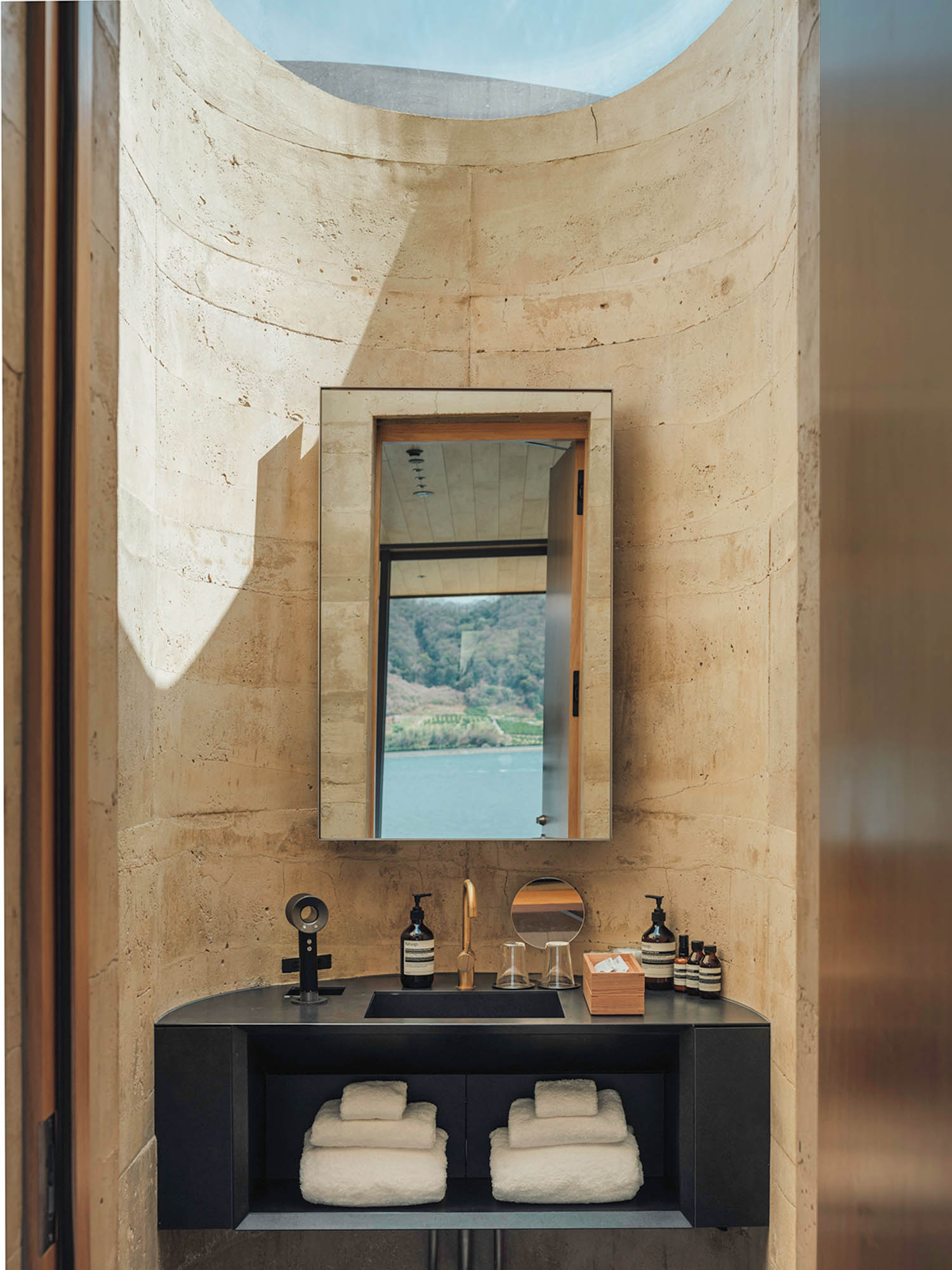 A bathroom vanity with a black sink, toiletries, and folded white towels, positioned against a curved stone wall under a skylight, with a mirror reflecting a view of a lake and hills.