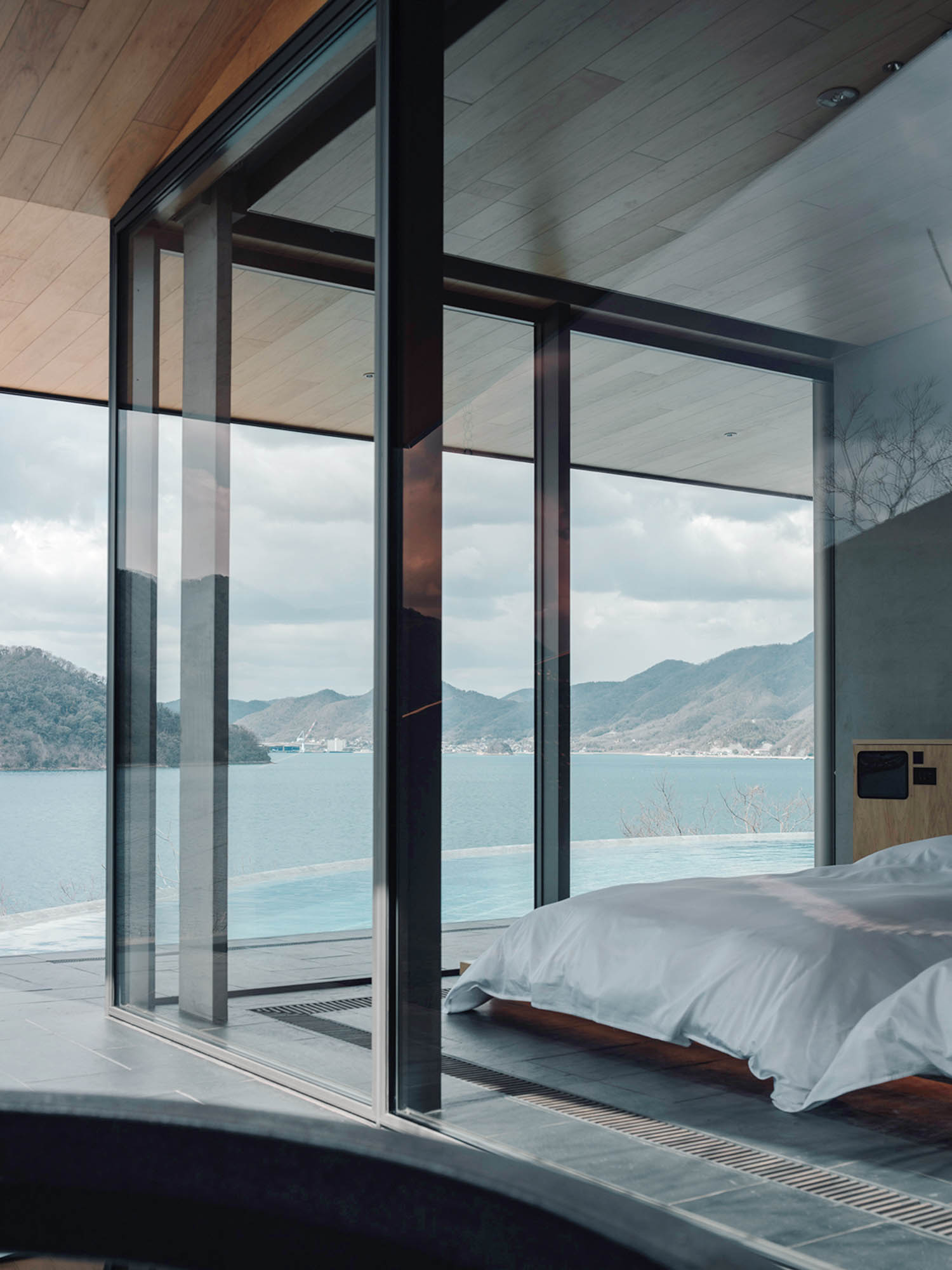 Modern bedroom with floor-to-ceiling glass walls overlooking a lake and mountain range.