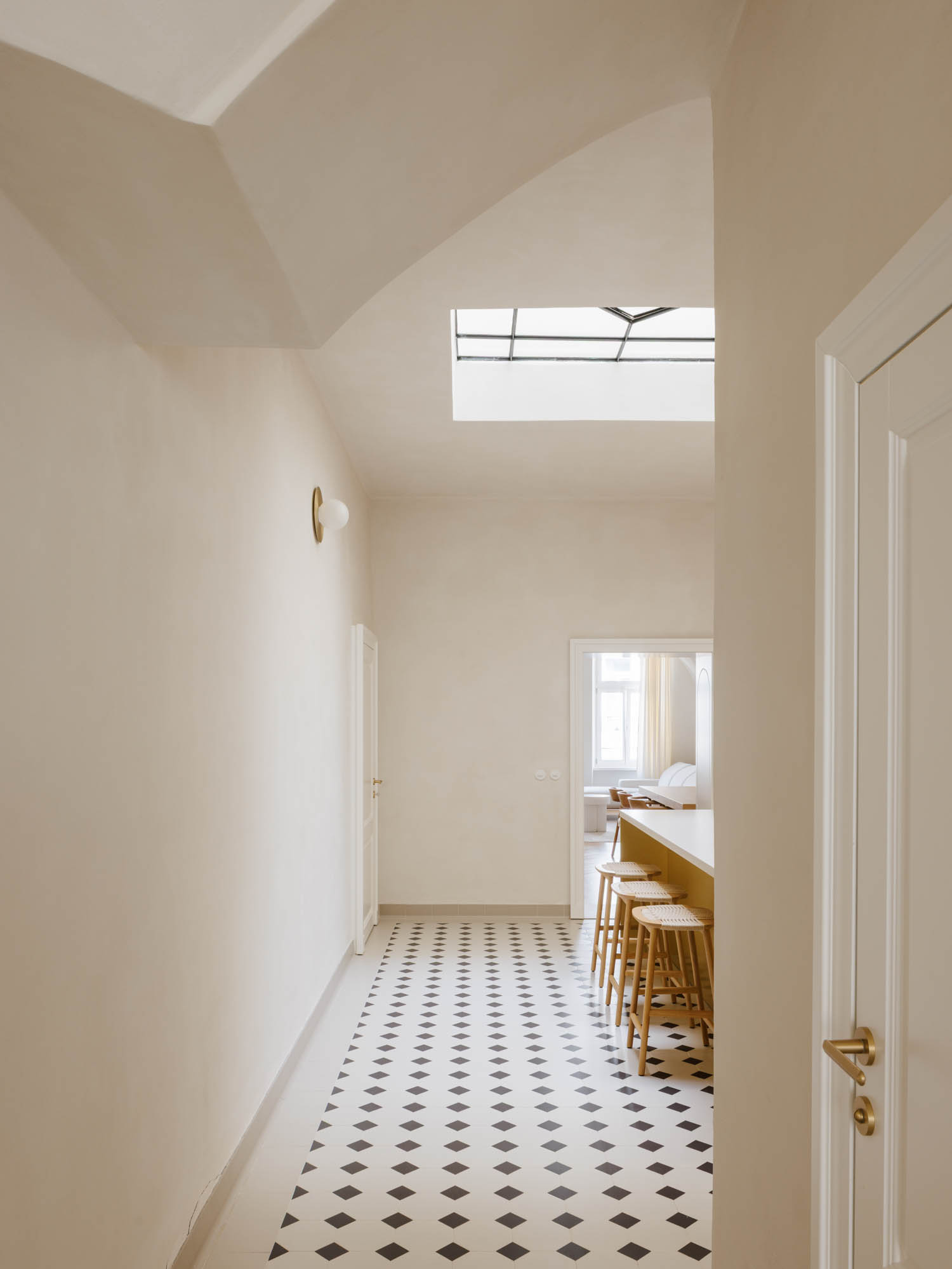 A hallway with a diamond-patterned tiled floor and a skylight, leading to a kitchen counter with three wooden bar stools.