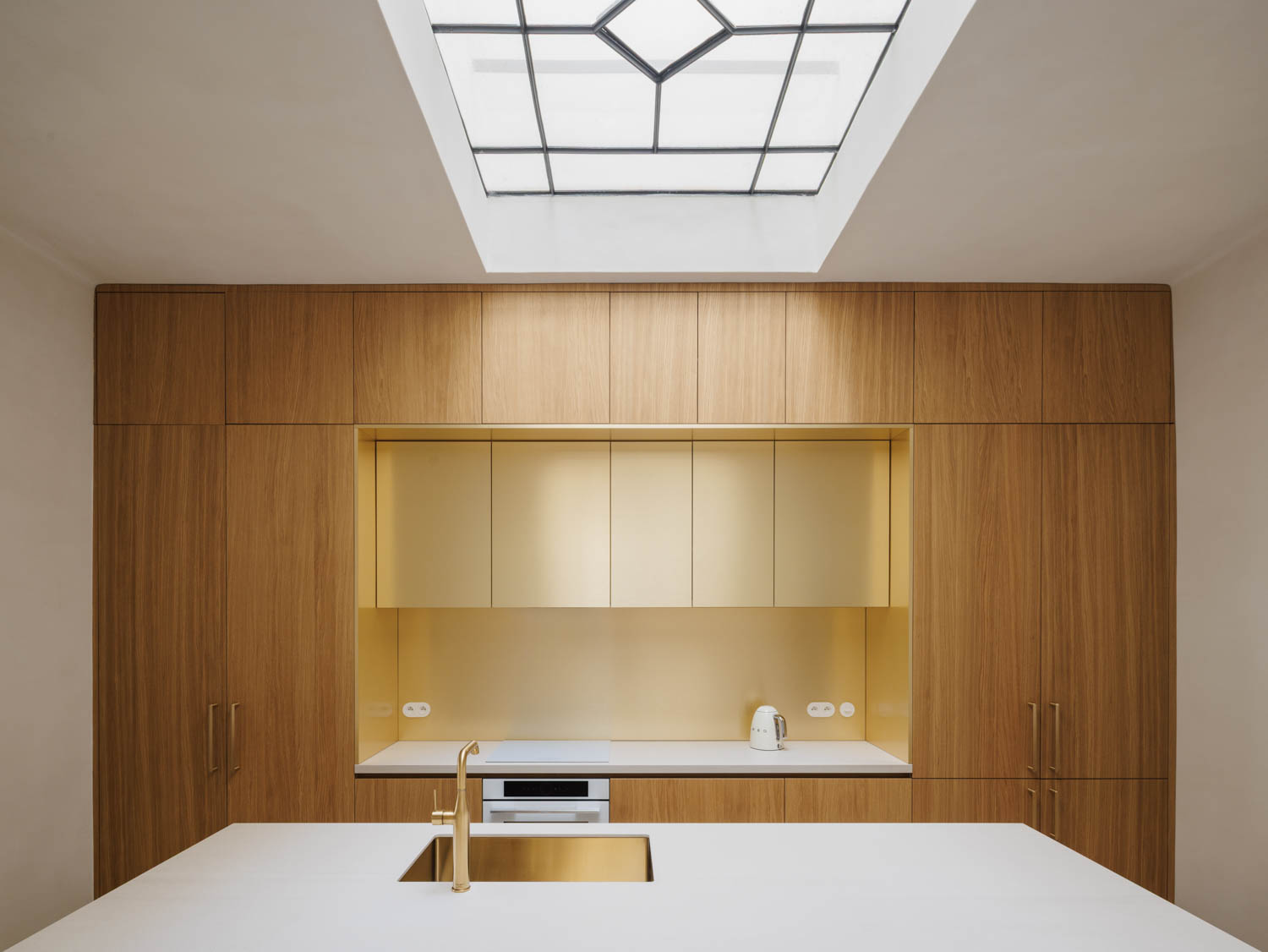 Modern kitchen with light wood cabinetry, gold-toned backsplash and sink, and a central island under a large skylight.