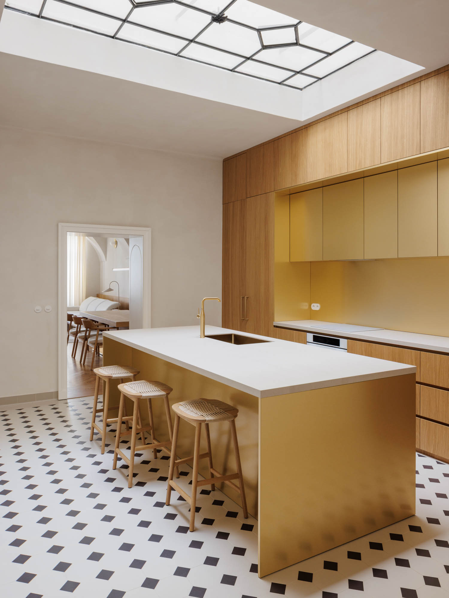 A kitchen island with a white countertop and metallic gold base, surrounded by three wooden stools, set against matching gold cabinetry under a large skylight.