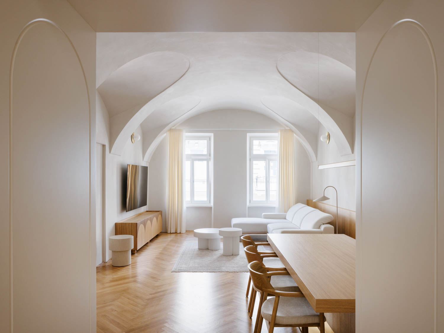 A living and dining room with a vaulted ceiling, herringbone wood floors, a white sectional sofa, and a long wooden dining table.