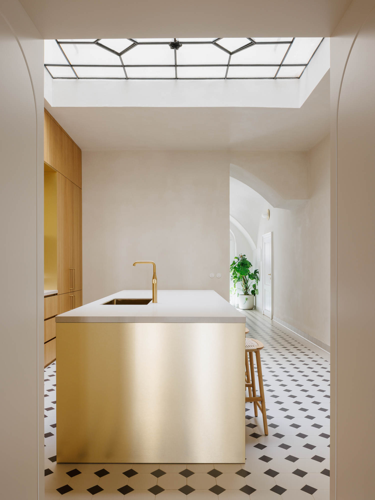 A kitchen island with a brass front and gold faucet sits in a room with a skylight, wood cabinetry, and a black-and-white checkered floor.