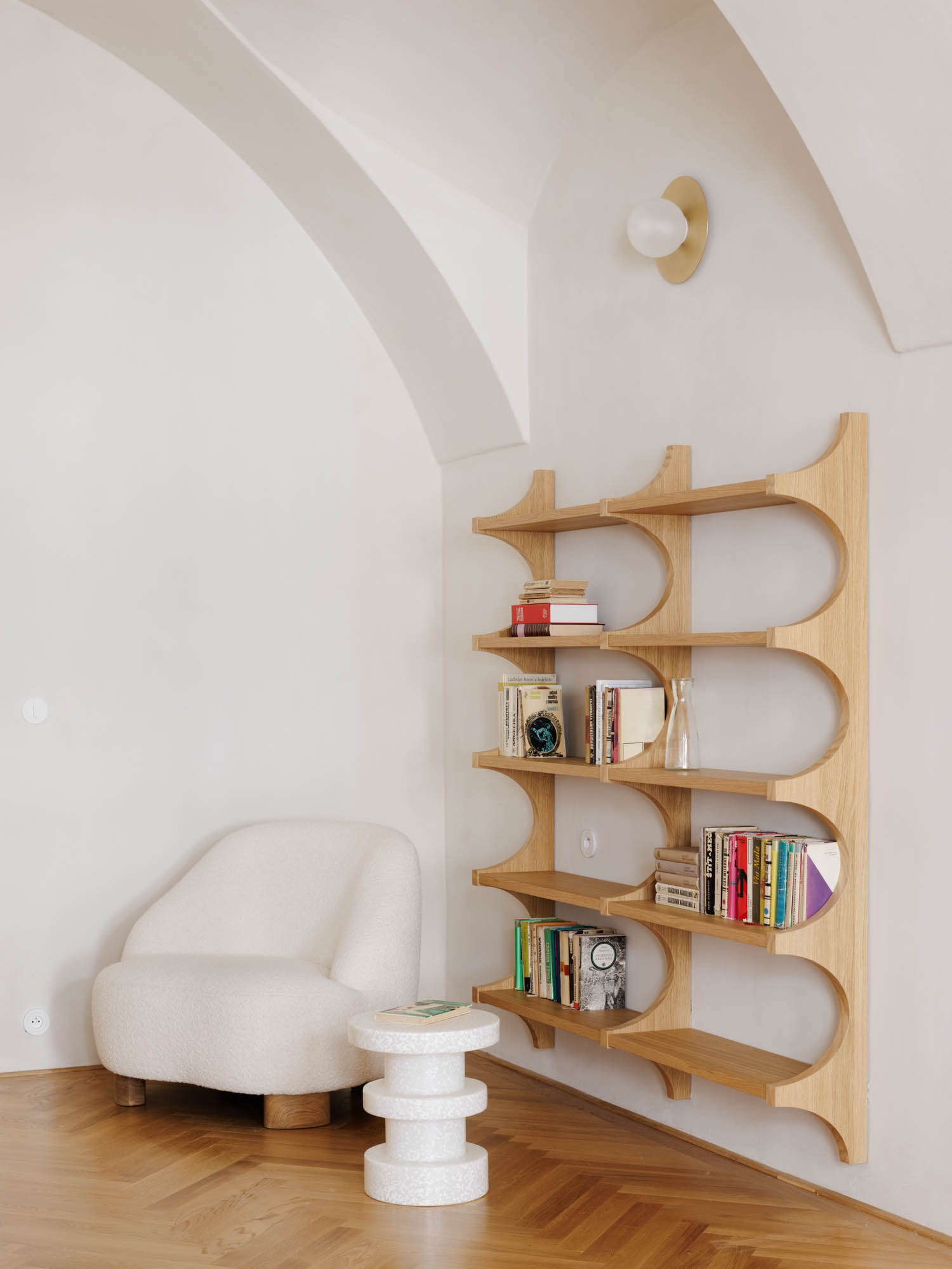 A white armchair and a small, cylindrical side table next to a large, wooden open-frame bookshelf against a white curved wall.